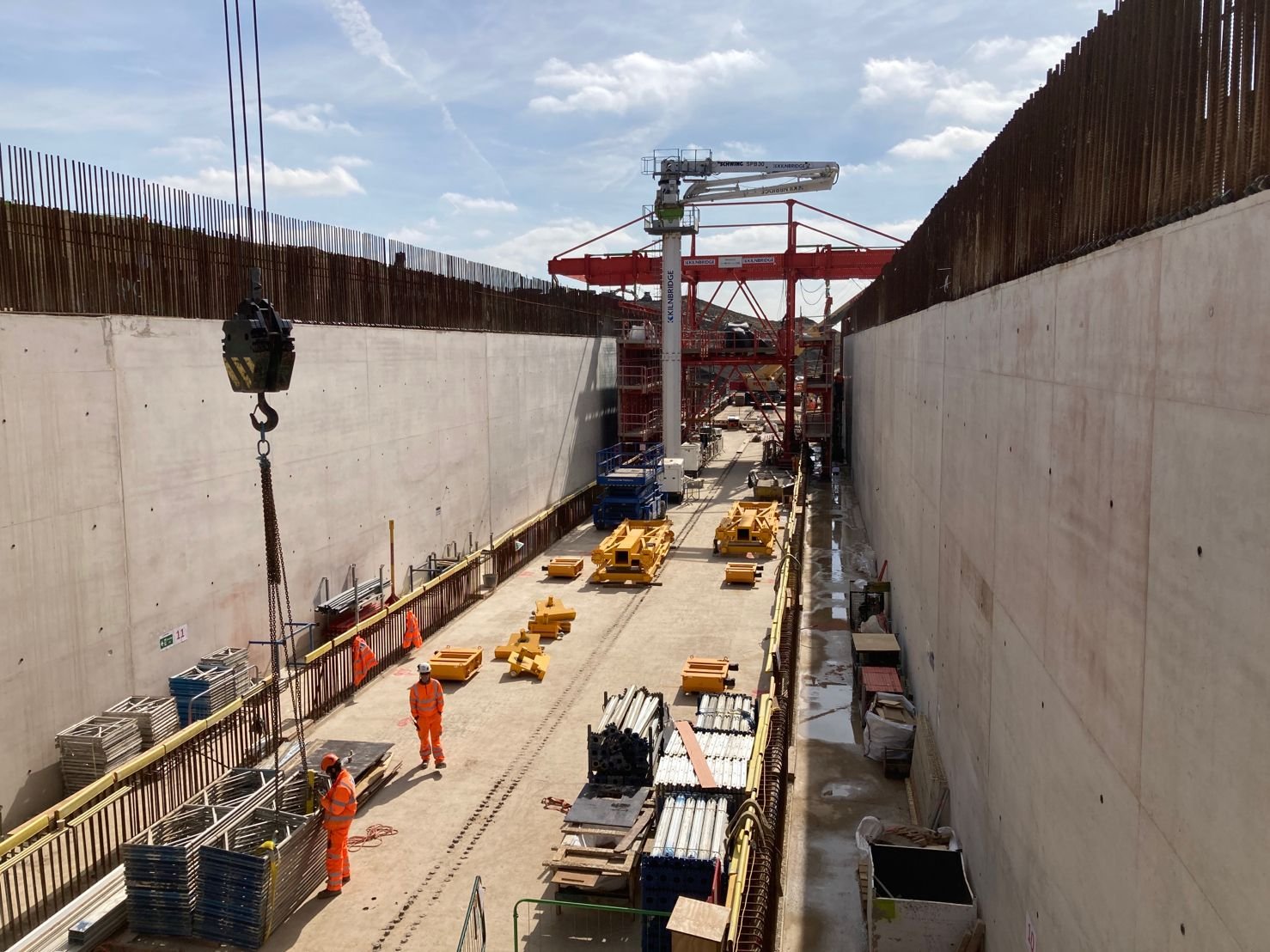 Construction site inside a large trench with workers in orange safety gear, construction equipment, and large red crane overhead.