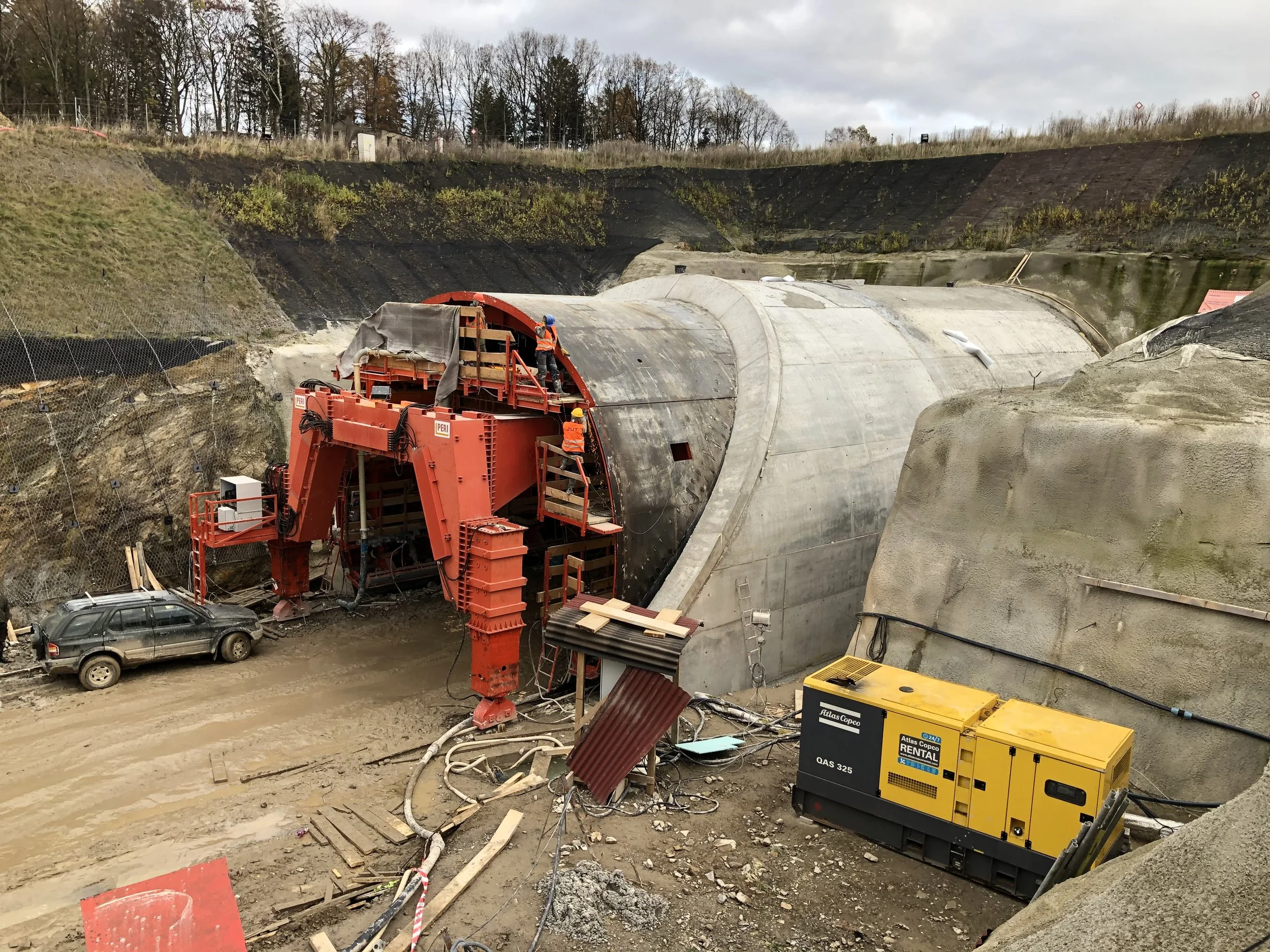 Construction workers installing a tunnel in a hillside surrounded by trees, with equipment like a large red tunnel boring machine and a yellow generator.
