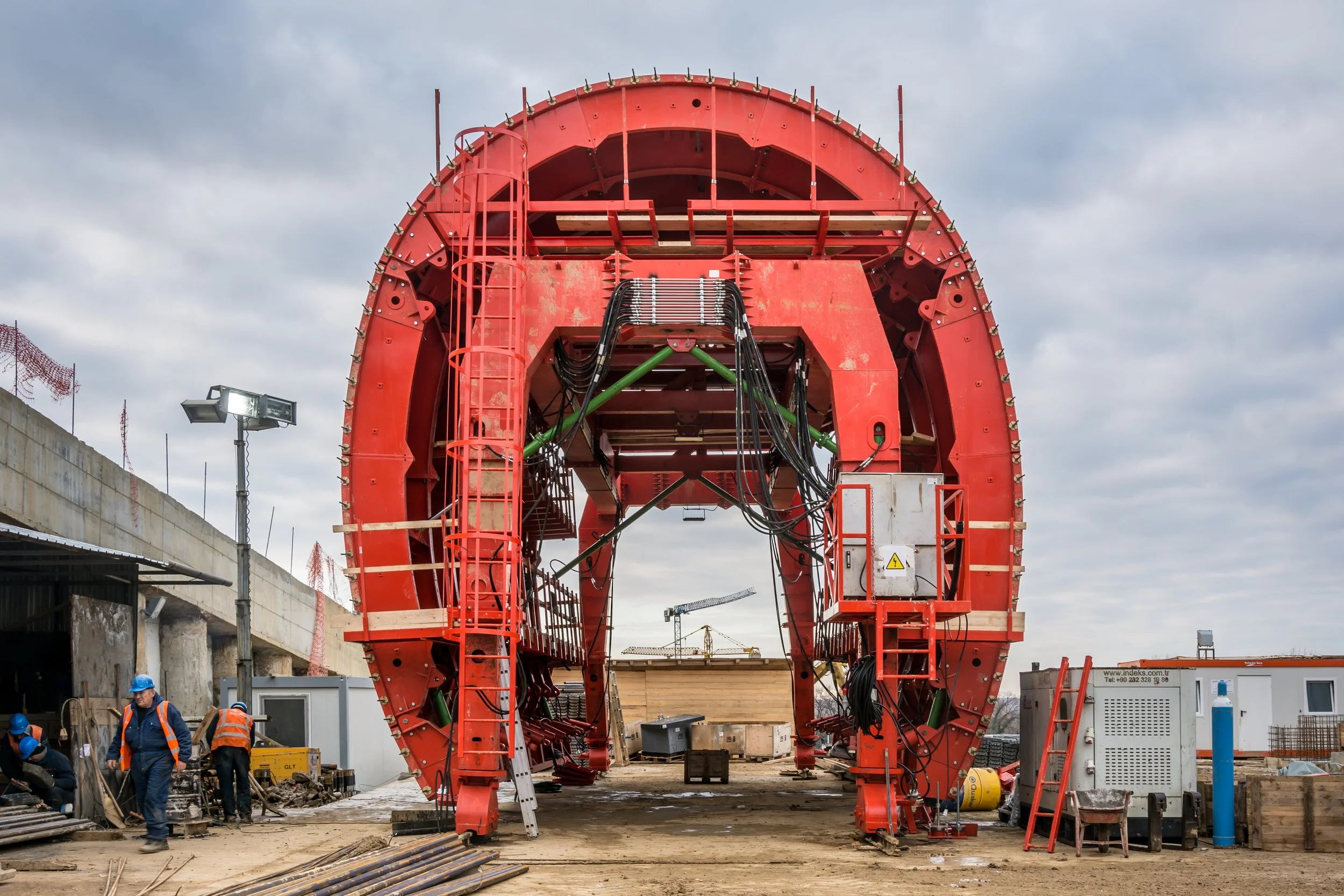 Construction workers in safety gear working near a large red tunnel boring machine on a construction site under cloudy skies.