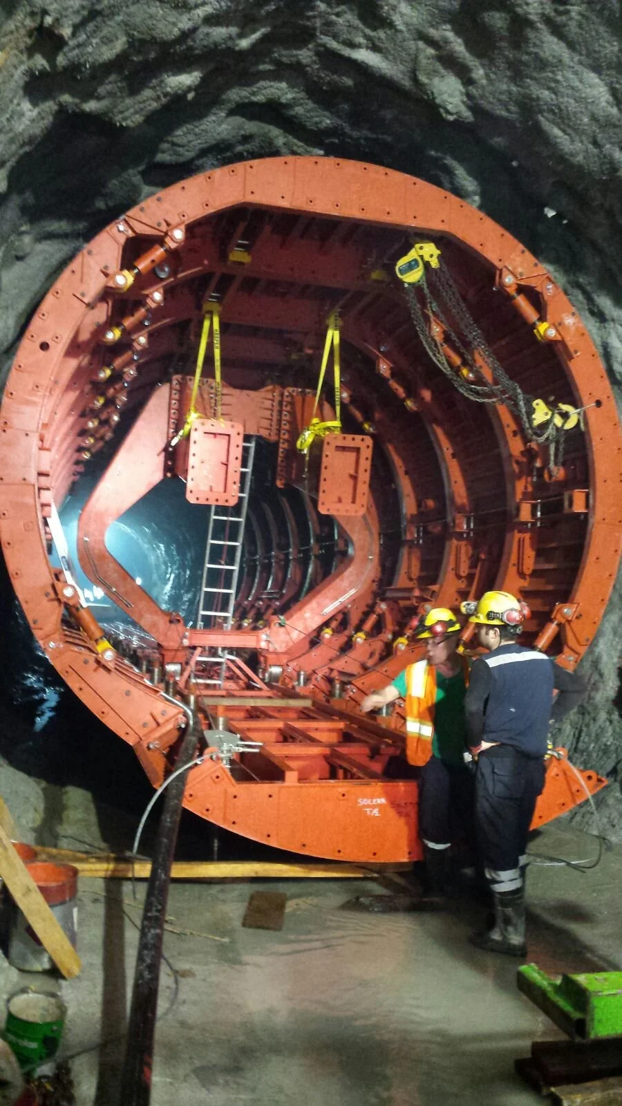 Workers in helmets and safety gear standing inside a large, cylindrical tunnel with orange lining, surrounded by rocky walls, engaged in construction or inspection.