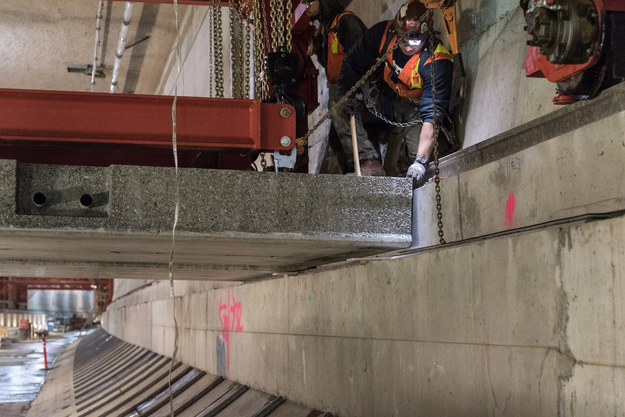 Construction workers in safety gear working on a concrete building installation, positioning a large concrete beam with chains and a hoist in an industrial setting.