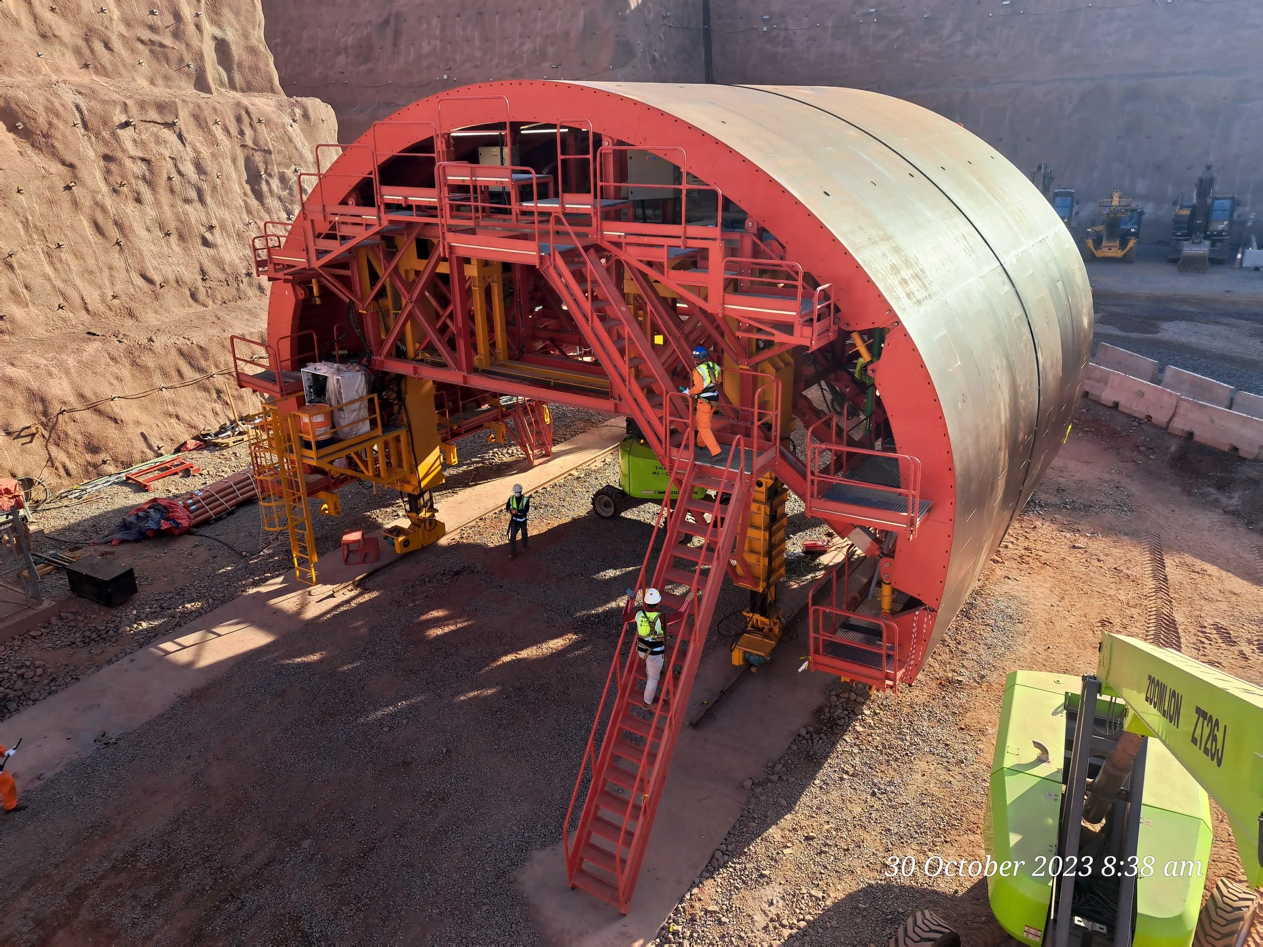 Construction site showing a large red and black tunnel boring machine with workers around it, and various construction equipment and machinery in the background.