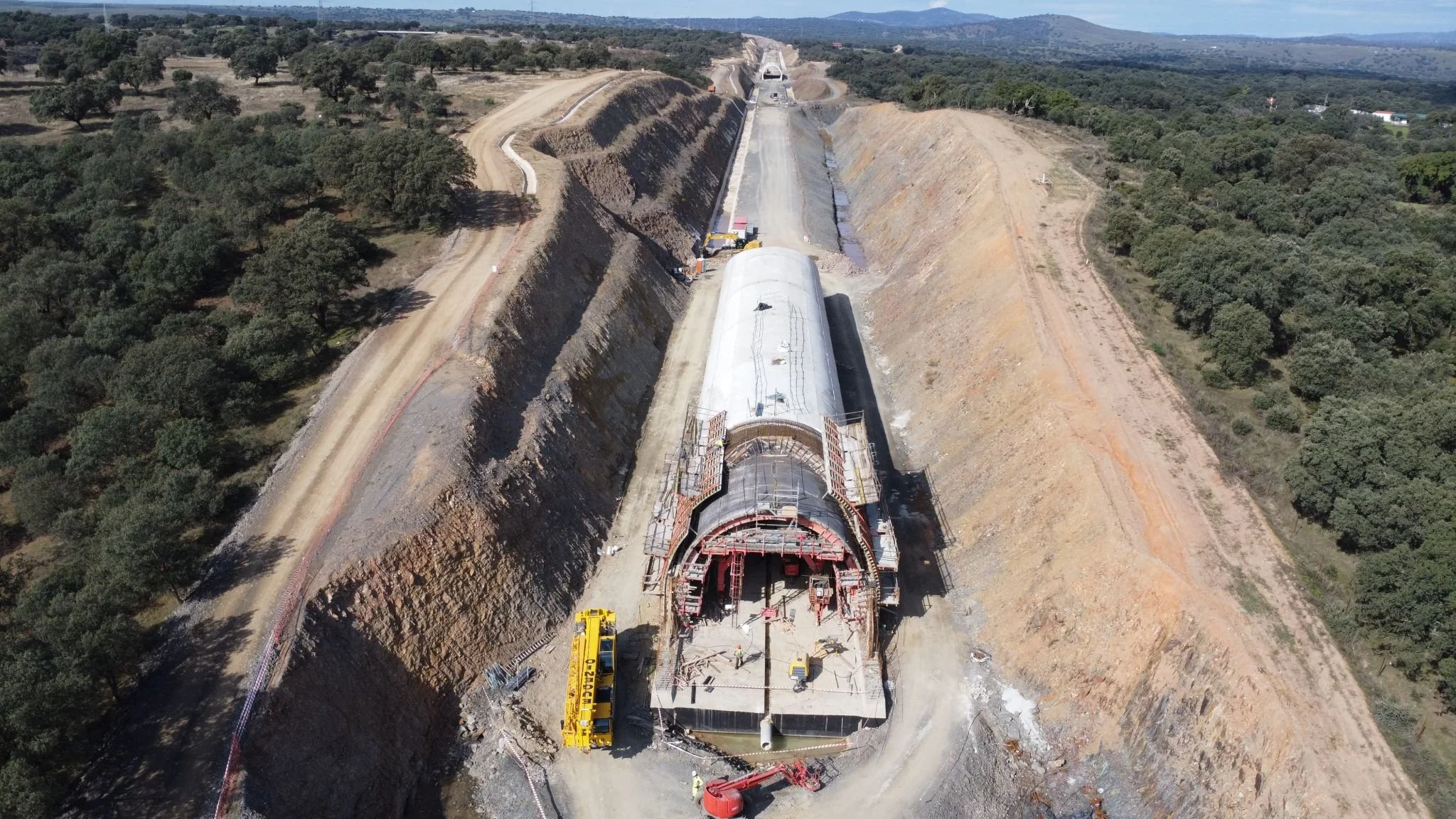 Aerial view of a construction site with a large tunnel boring machine in the process of excavation, surrounded by hills and trees, with ongoing road construction and machinery present.