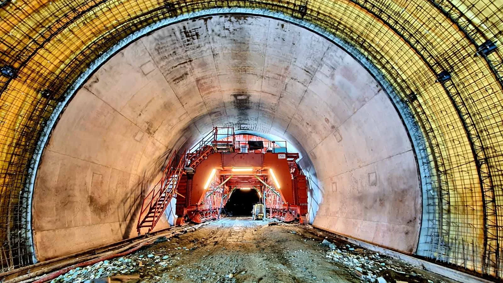 View inside a large tunnel under construction, showing concrete arch with yellow safety netting and a red machine at the far end.