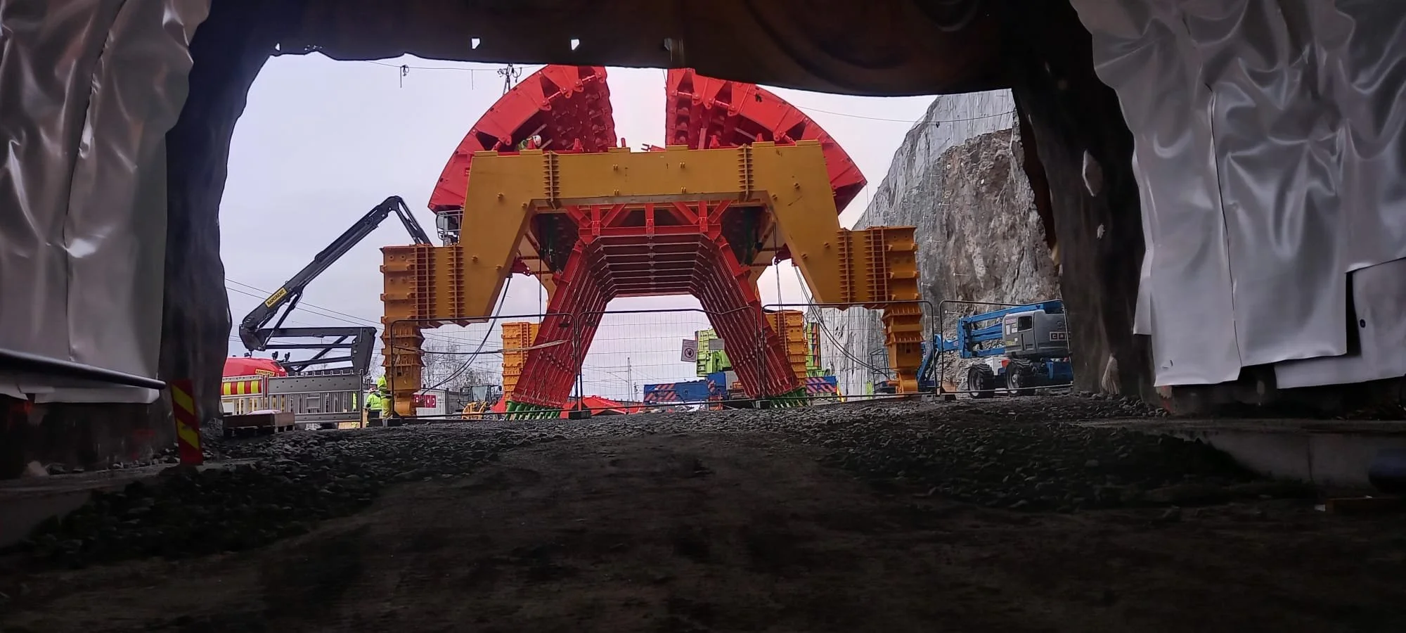 View from inside a tunnel showing construction of a large red and yellow tunnel boring machine (TBM) emerging at the exit, with construction workers and equipment nearby.