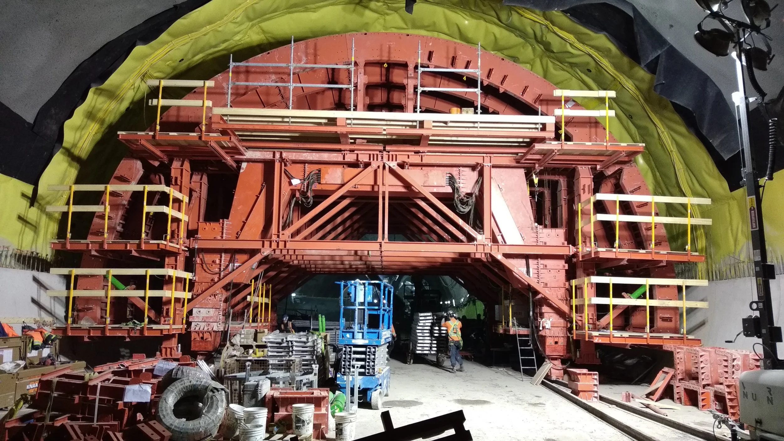 Construction workers and equipment inside a tunnel under construction, with scaffolding and structural elements visible.