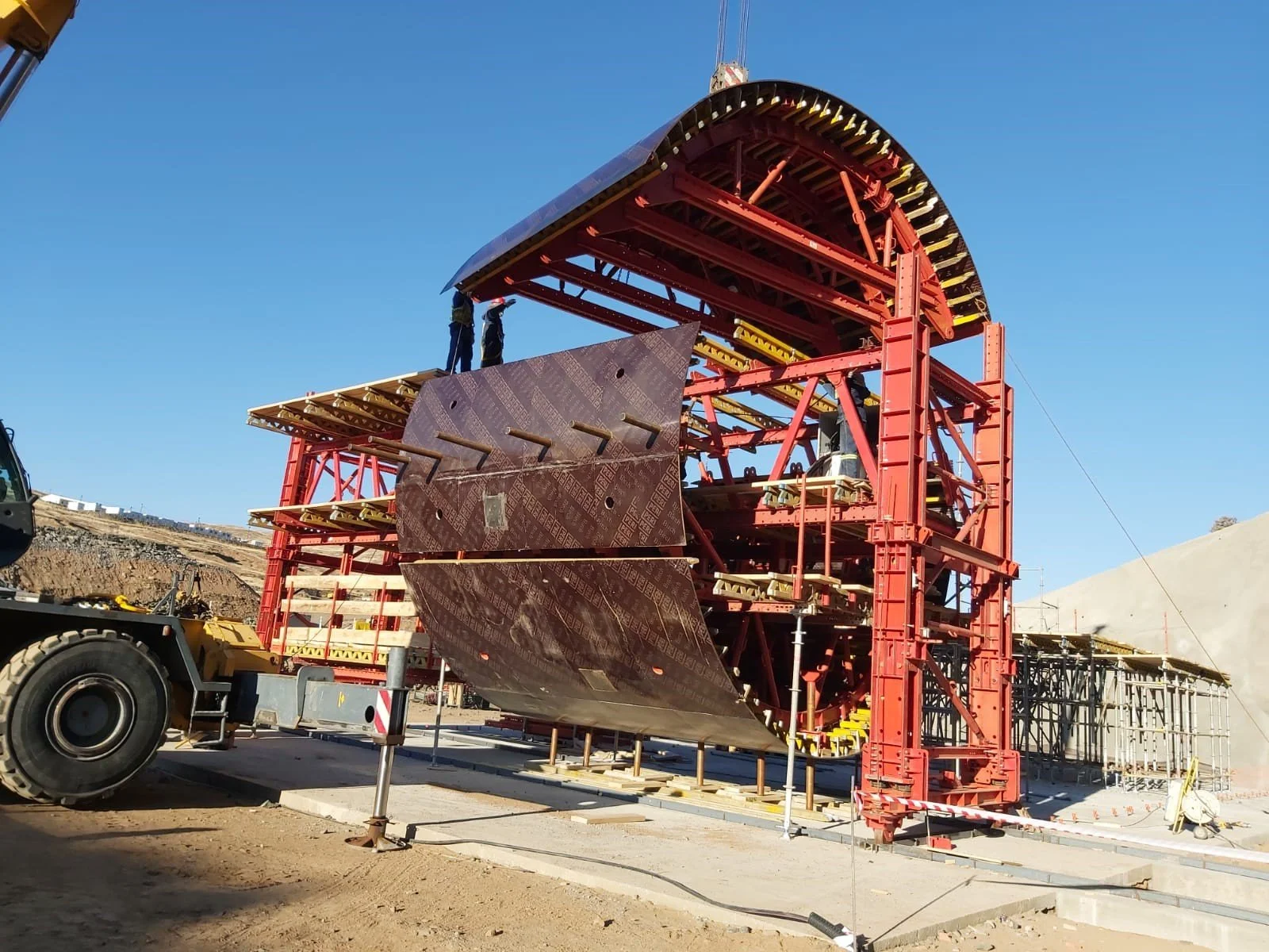 Construction site with a large red and black metal structure, workers on top, scaffolding and construction equipment visible, clear blue sky.