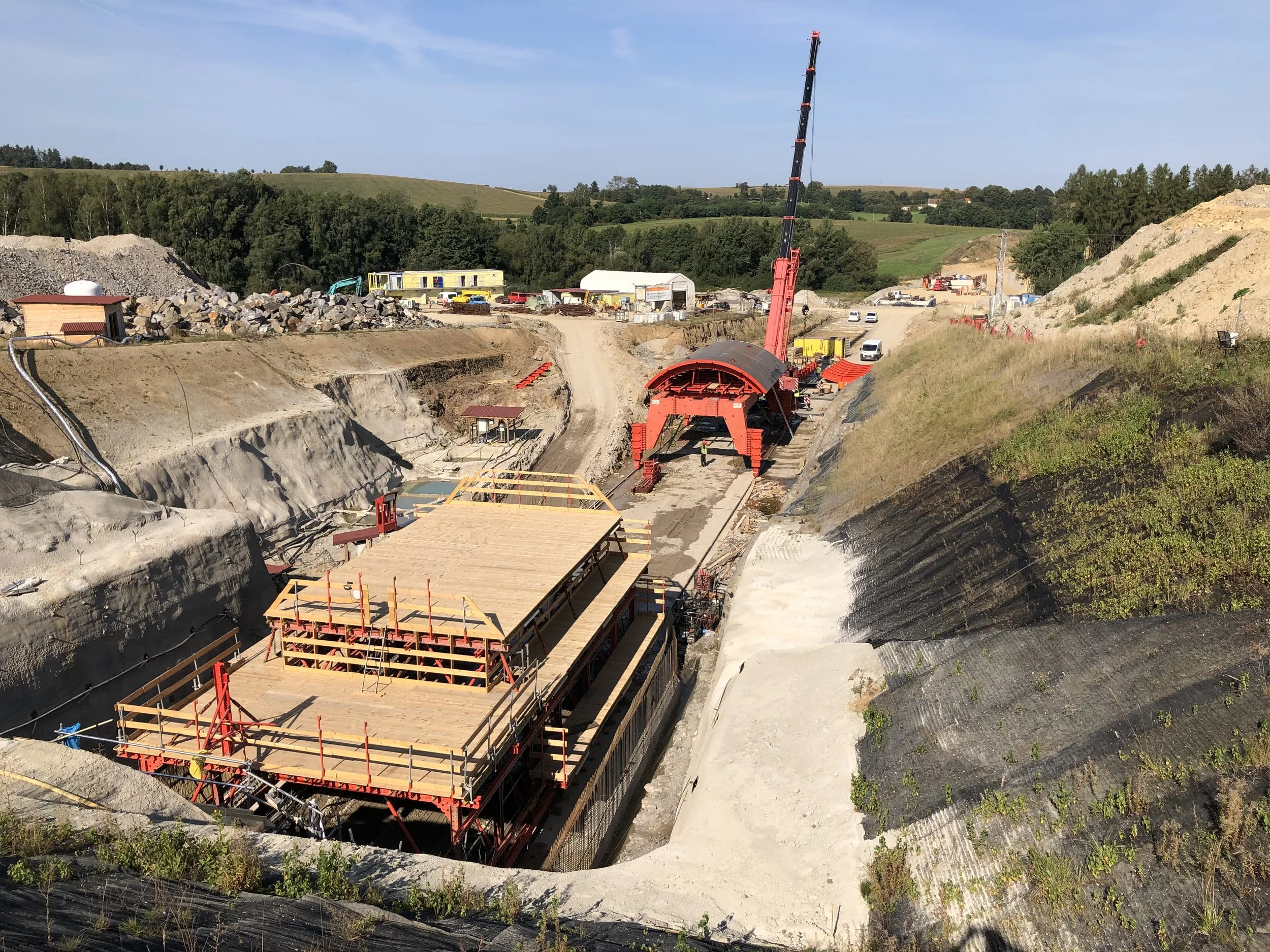 Construction site with large red crane, wooden platform, and excavated hillside under clear blue sky.