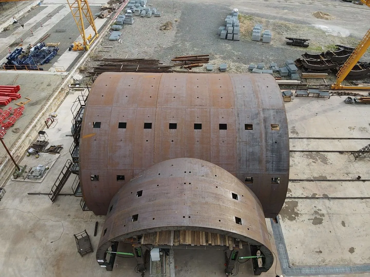 Construction site with two large, rusted, curved steel structures, various building materials, equipment, and yellow cranes.