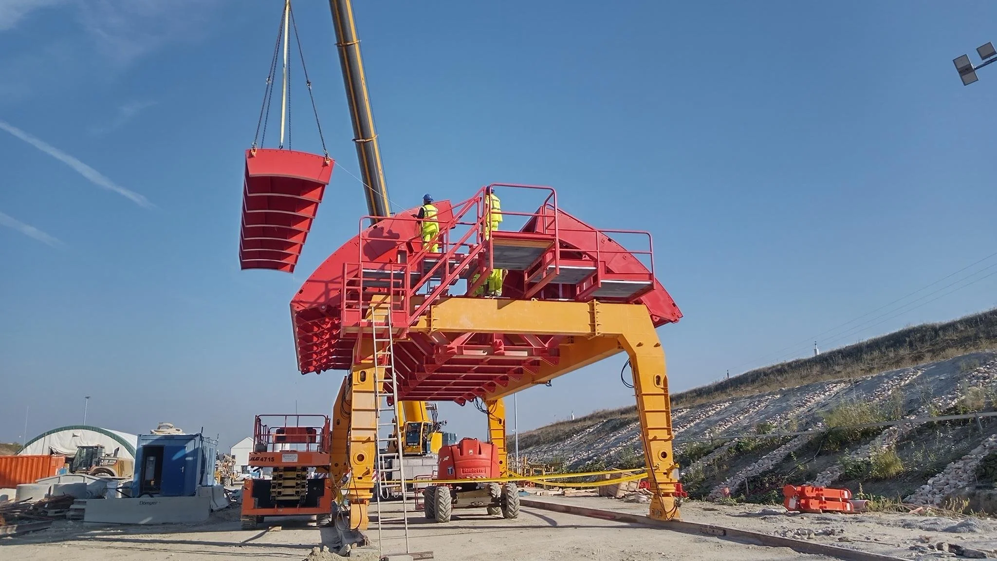 Construction workers in yellow safety vests and helmets working on a large robotic machine with red and yellow parts at a construction site under a clear blue sky.