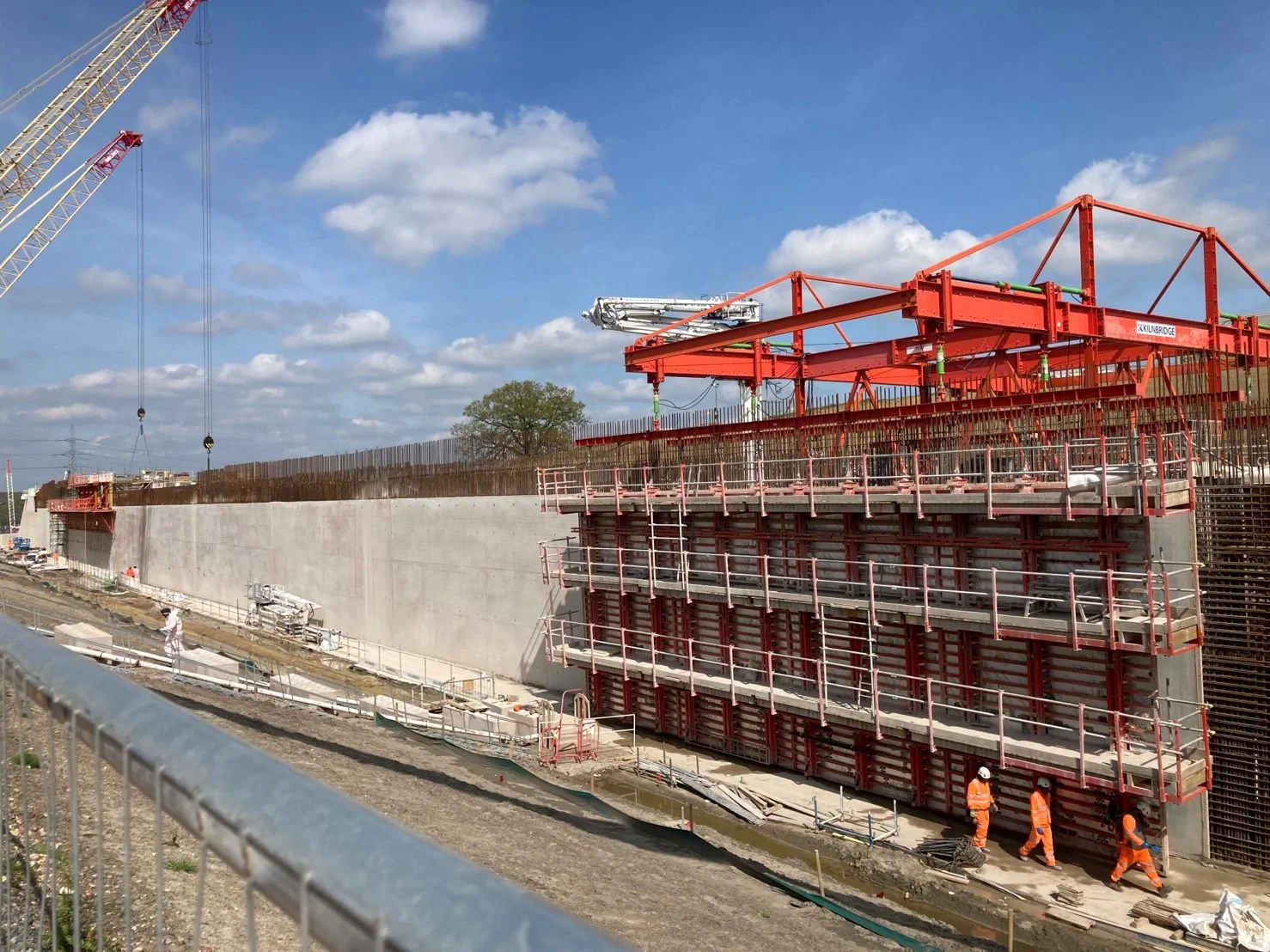 Construction site with a large concrete wall, scaffolding, and workers in orange safety uniforms, under a partly cloudy sky.