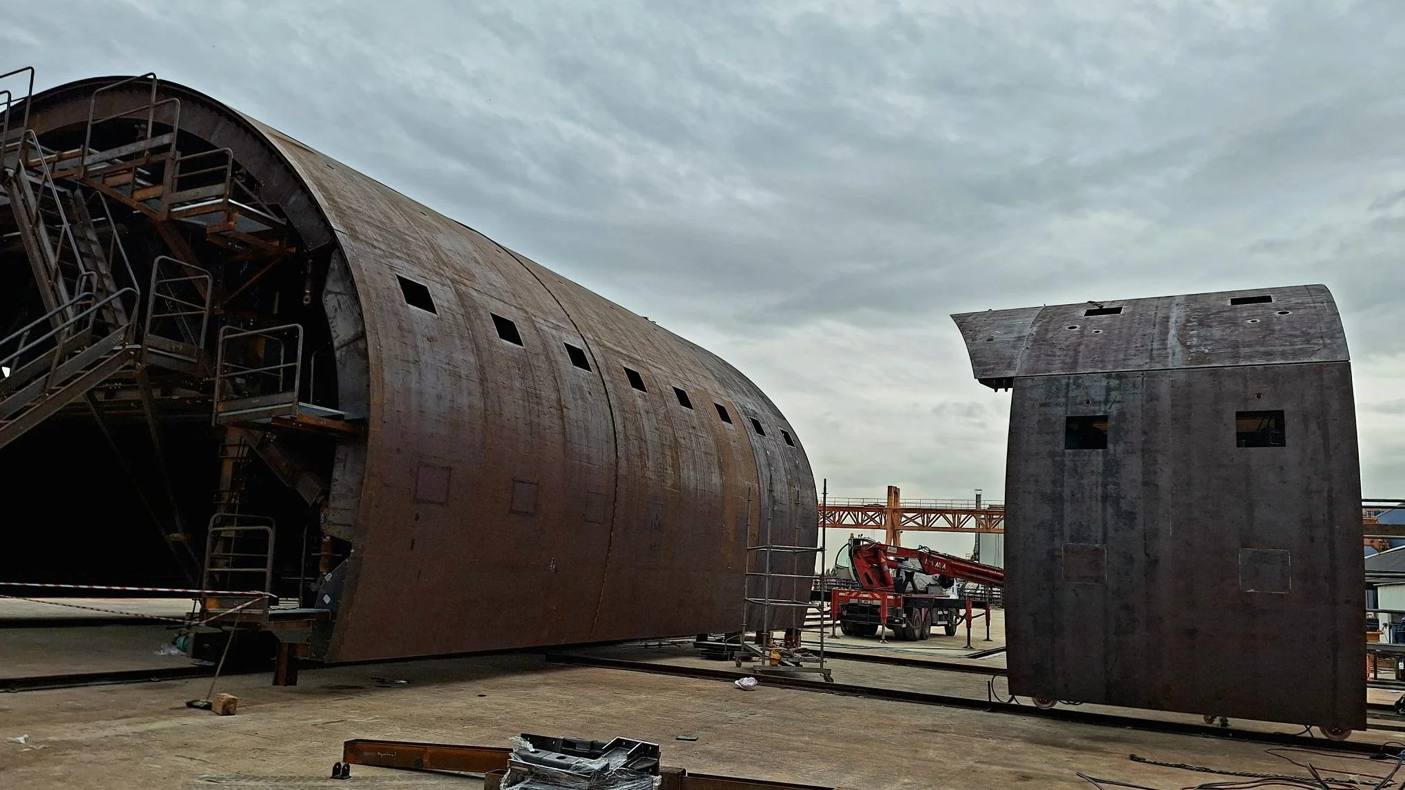 Large unfinished metal structure with multiple small windows, scaffolding, and a crane in the background at a construction site