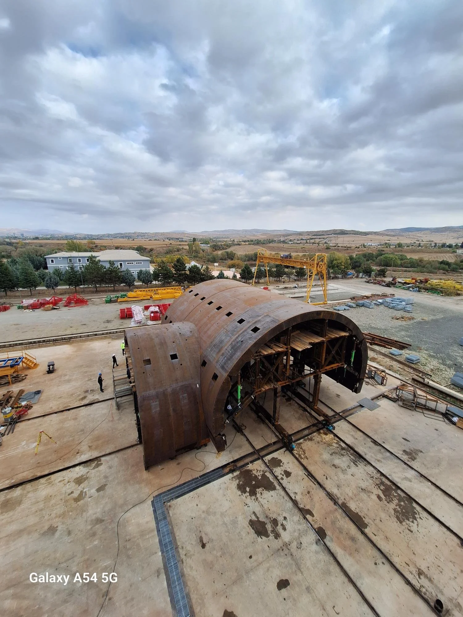Construction site with large, curved steel structure under development, surrounded by equipment, workers, and tracks, with a rural landscape and cloudy sky in the background.