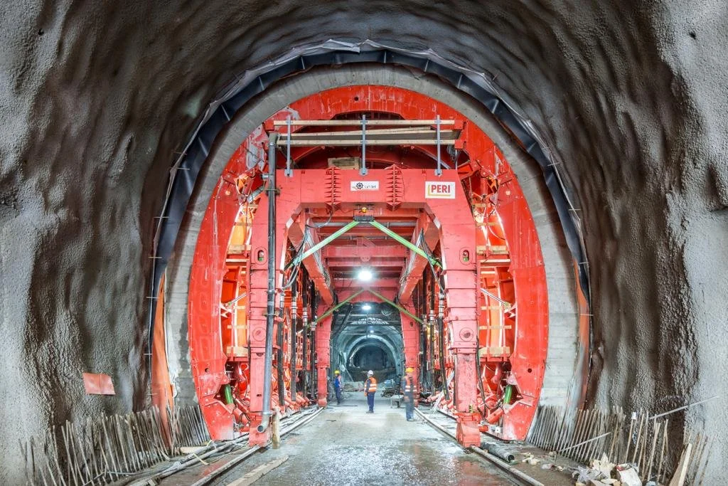 Construction workers inside a tunnel under construction, with large red tunnel boring machine components and excavation equipment.