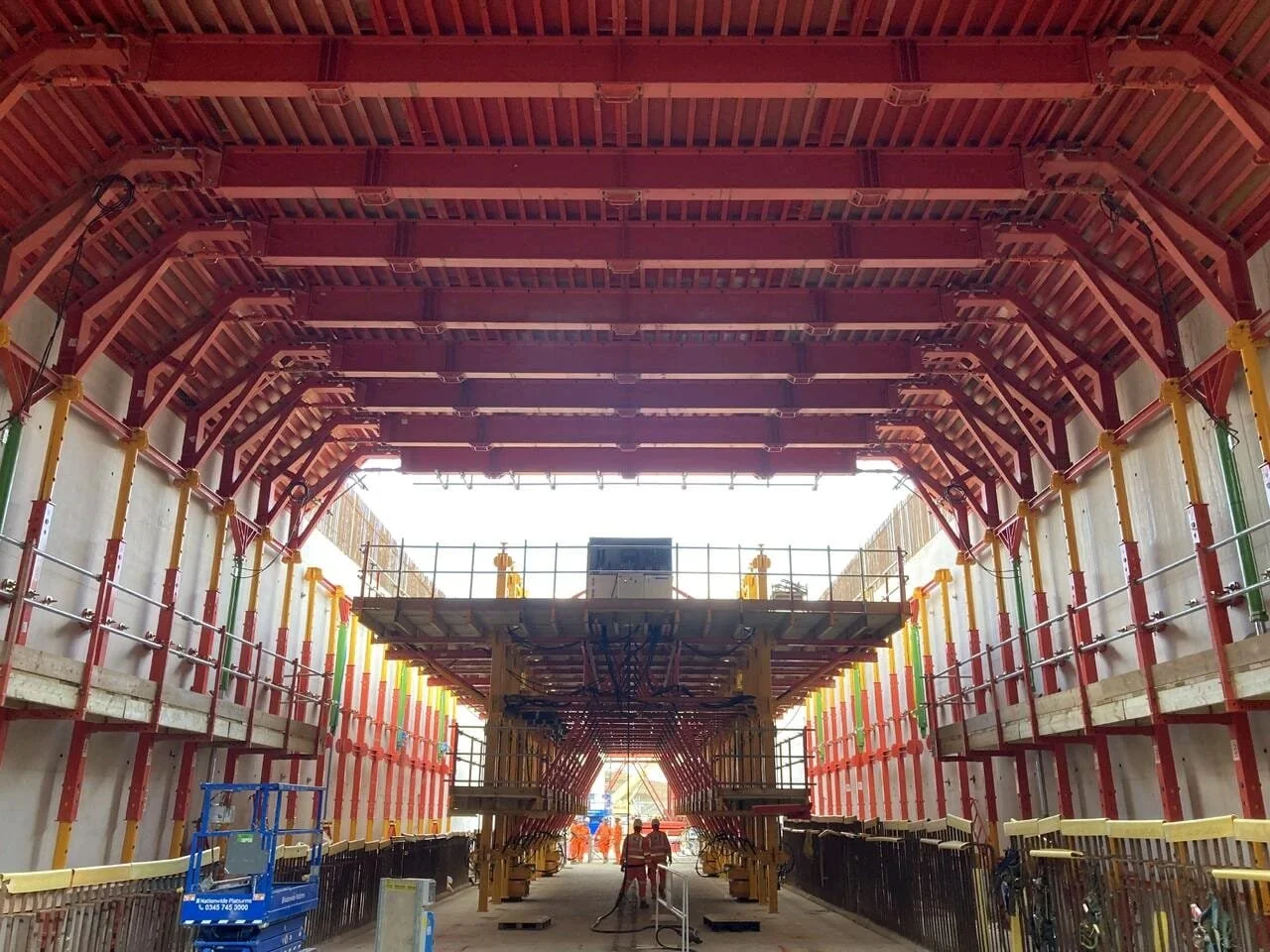 Construction site inside a building with a large arched wooden ceiling, scaffolding, and workers in orange safety vests.