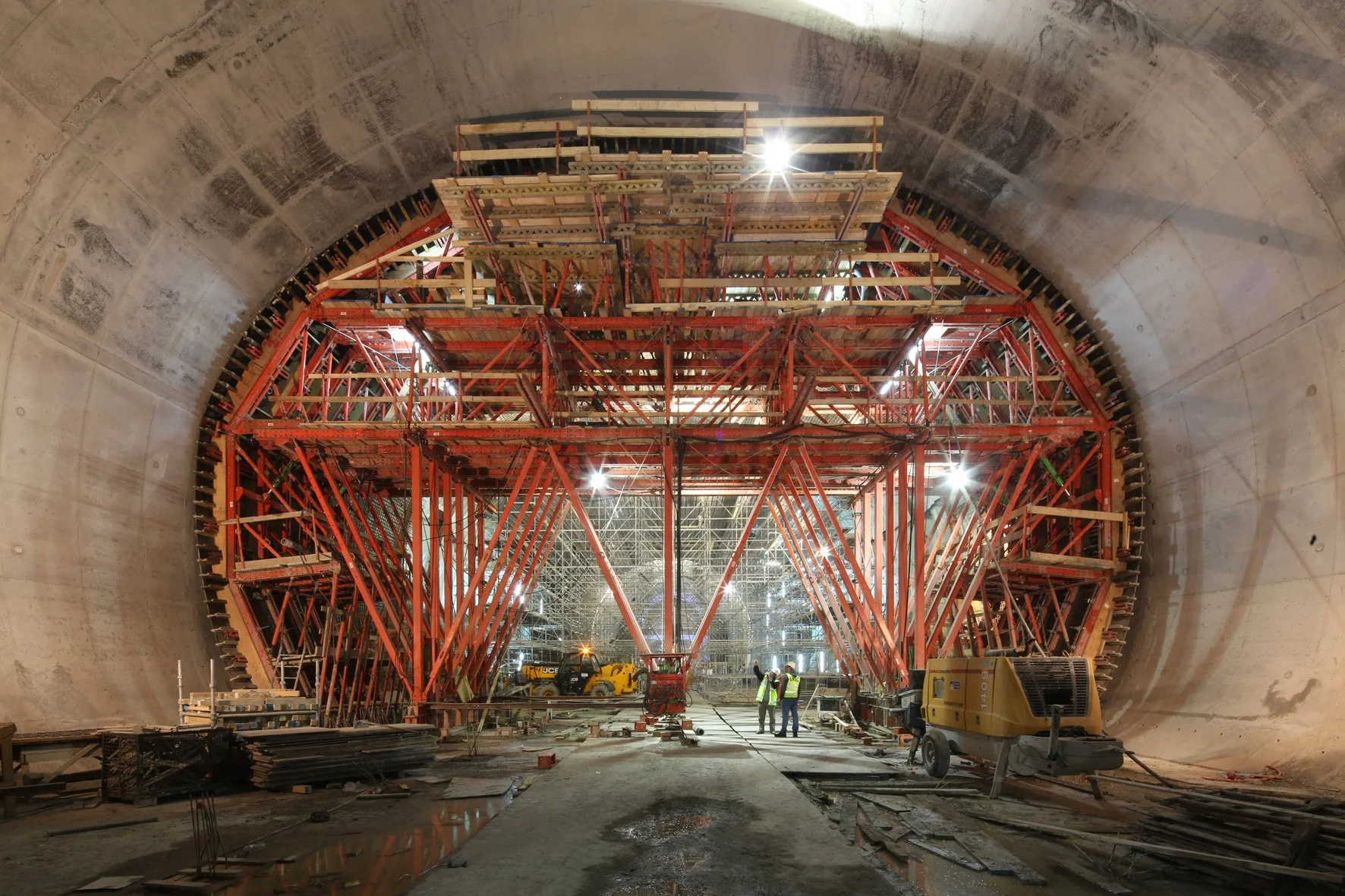 Construction workers and heavy machinery inside a large tunnel under construction, with metal scaffolding and support beams.