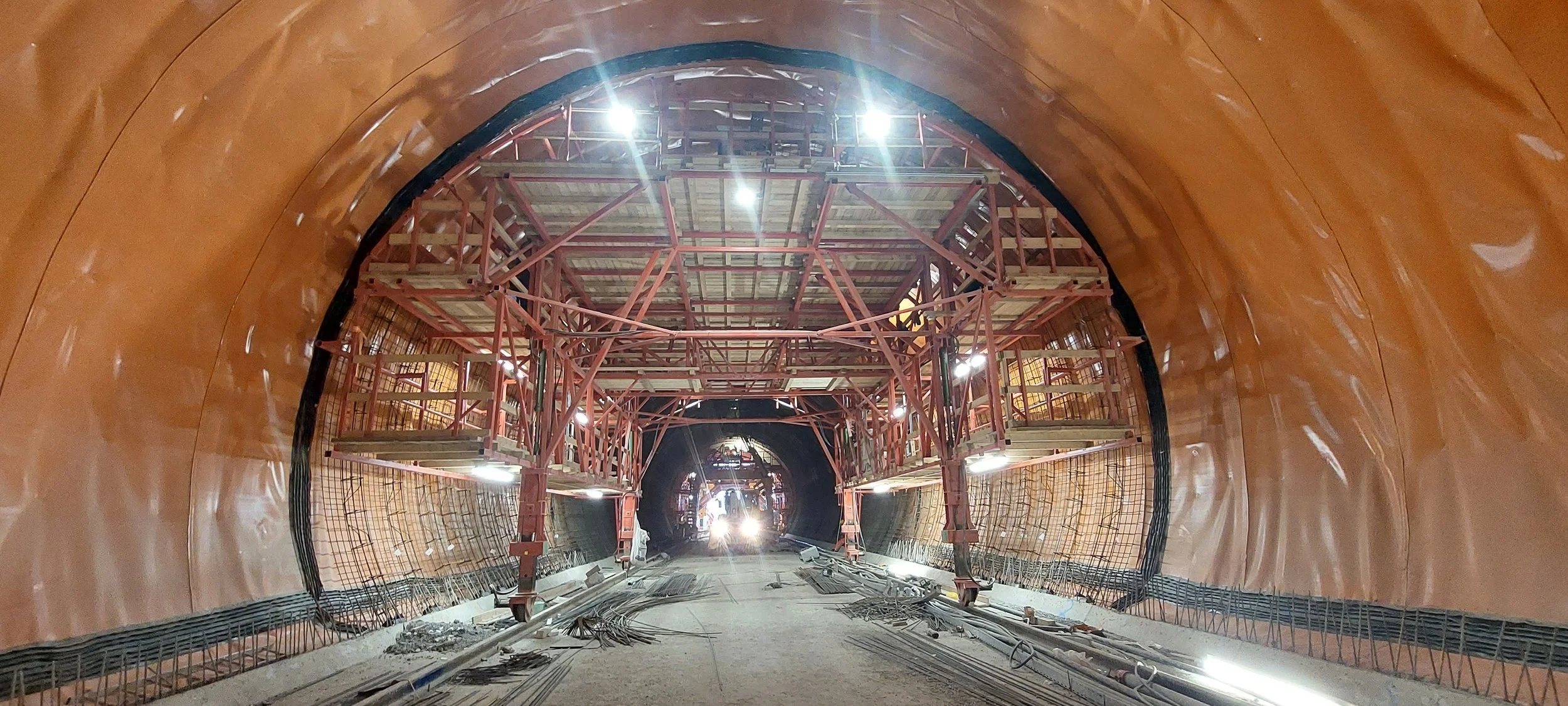 Under construction tunnel with orange lining, scaffolding and construction materials in the tunnel.