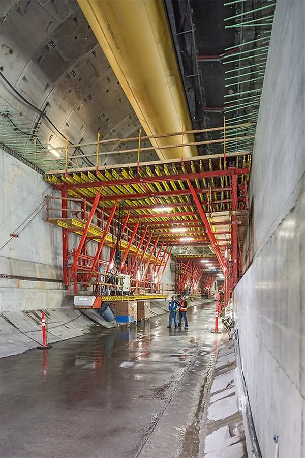 Construction workers inspecting inside a flood-prepared tunnel with large yellow and black pipes and red scaffolding.