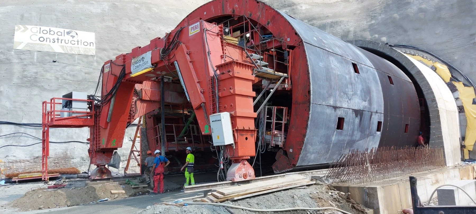 Construction workers and machinery at a tunnel construction site, with a large tunnel boring machine embedded in the ground.
