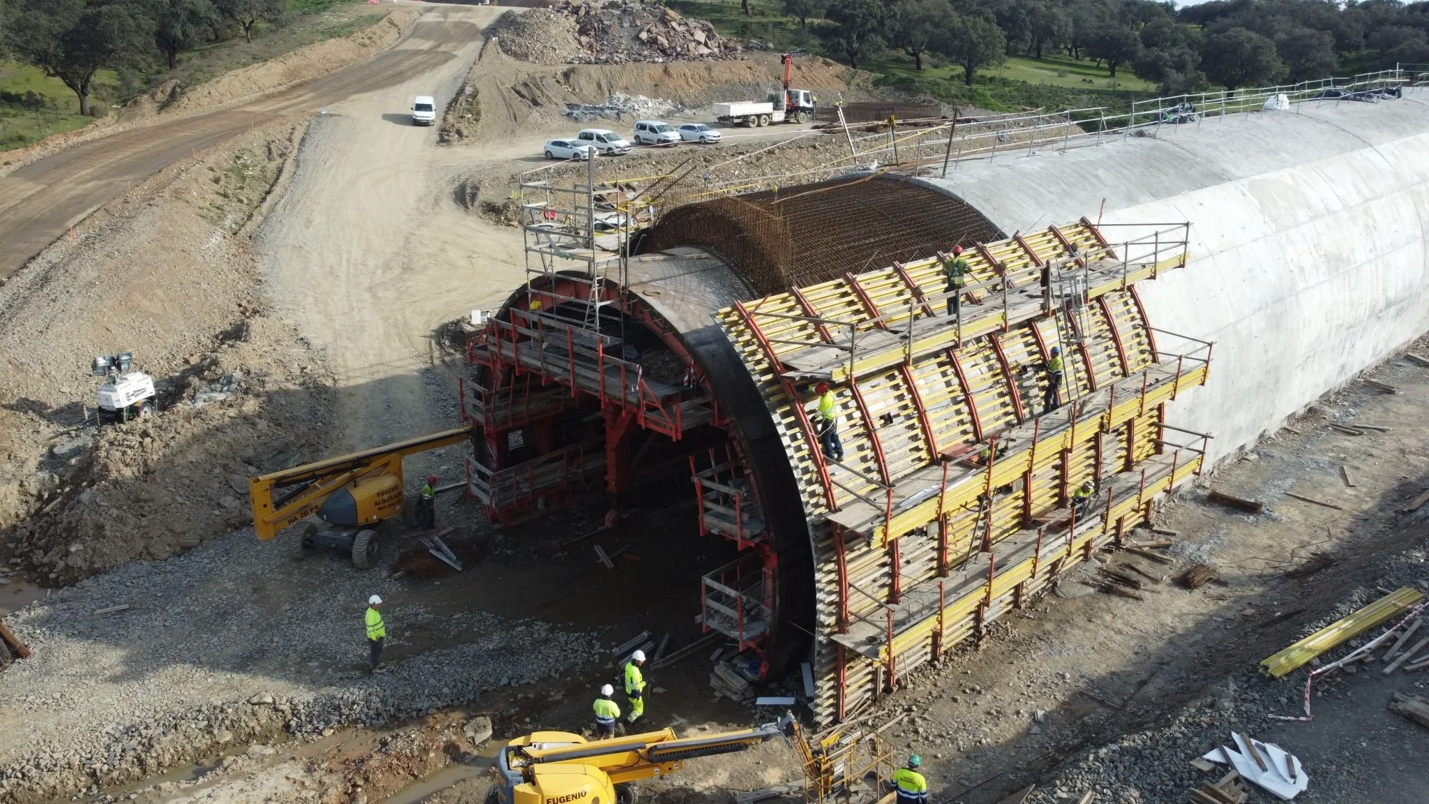 Construction workers install a large tunnel segment with scaffoldings, machinery, and construction materials around a tunnel entrance, with cars parked in the background on a dirt road.
