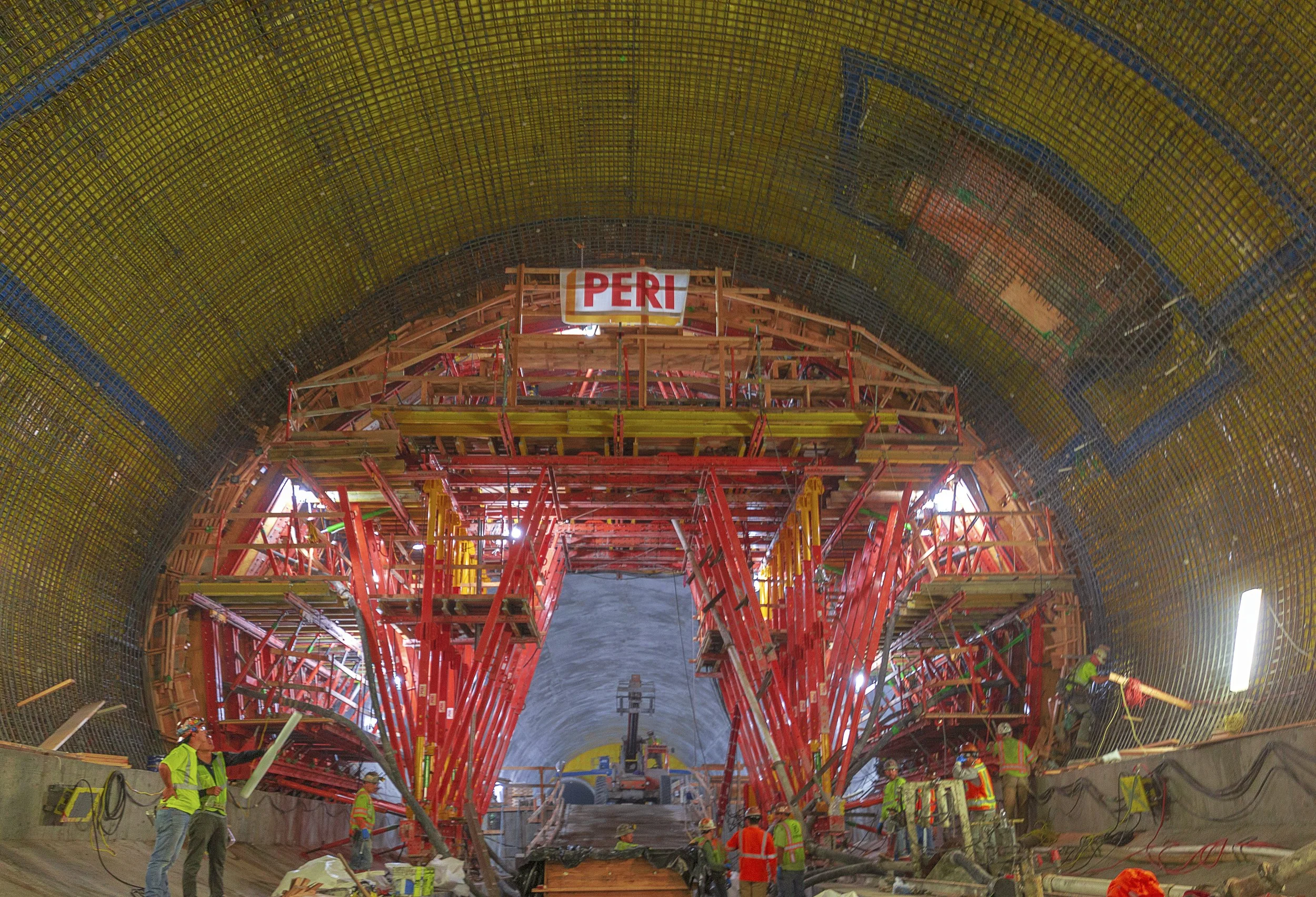 Interior of a tunnel construction site with workers in safety gear, scaffolding, and support structures, and a sign with the word 'PERU' at the top.
