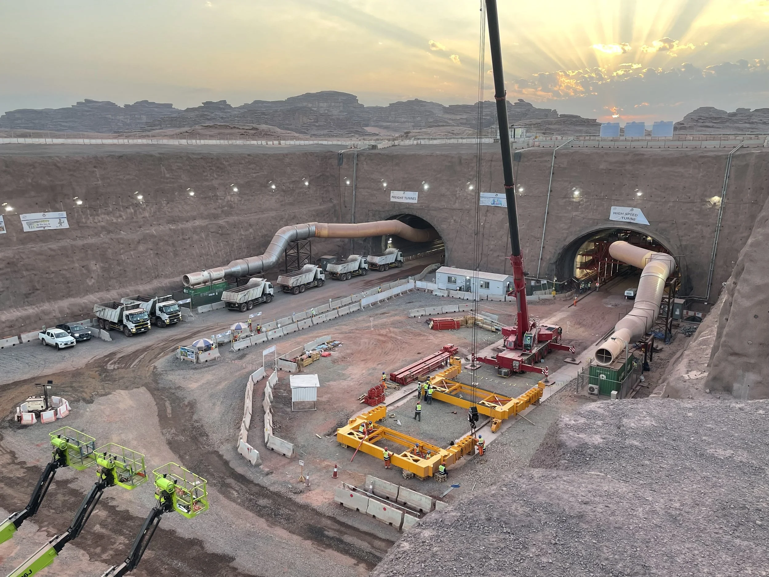 Construction workers and heavy machinery building tunnel underground in a desert landscape at sunset.