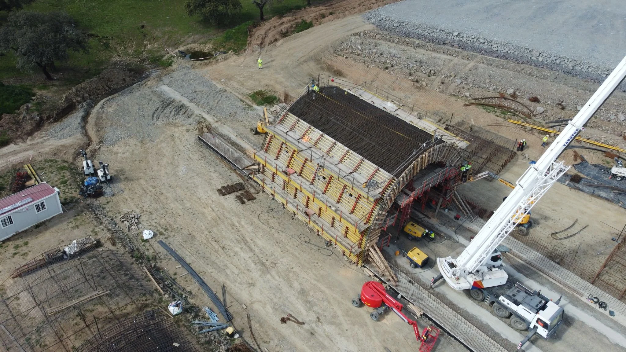 An aerial view of a construction site where workers are building a bridge with scaffolding and reinforcements. A large crane and construction vehicles are present, with materials and equipment scattered around.