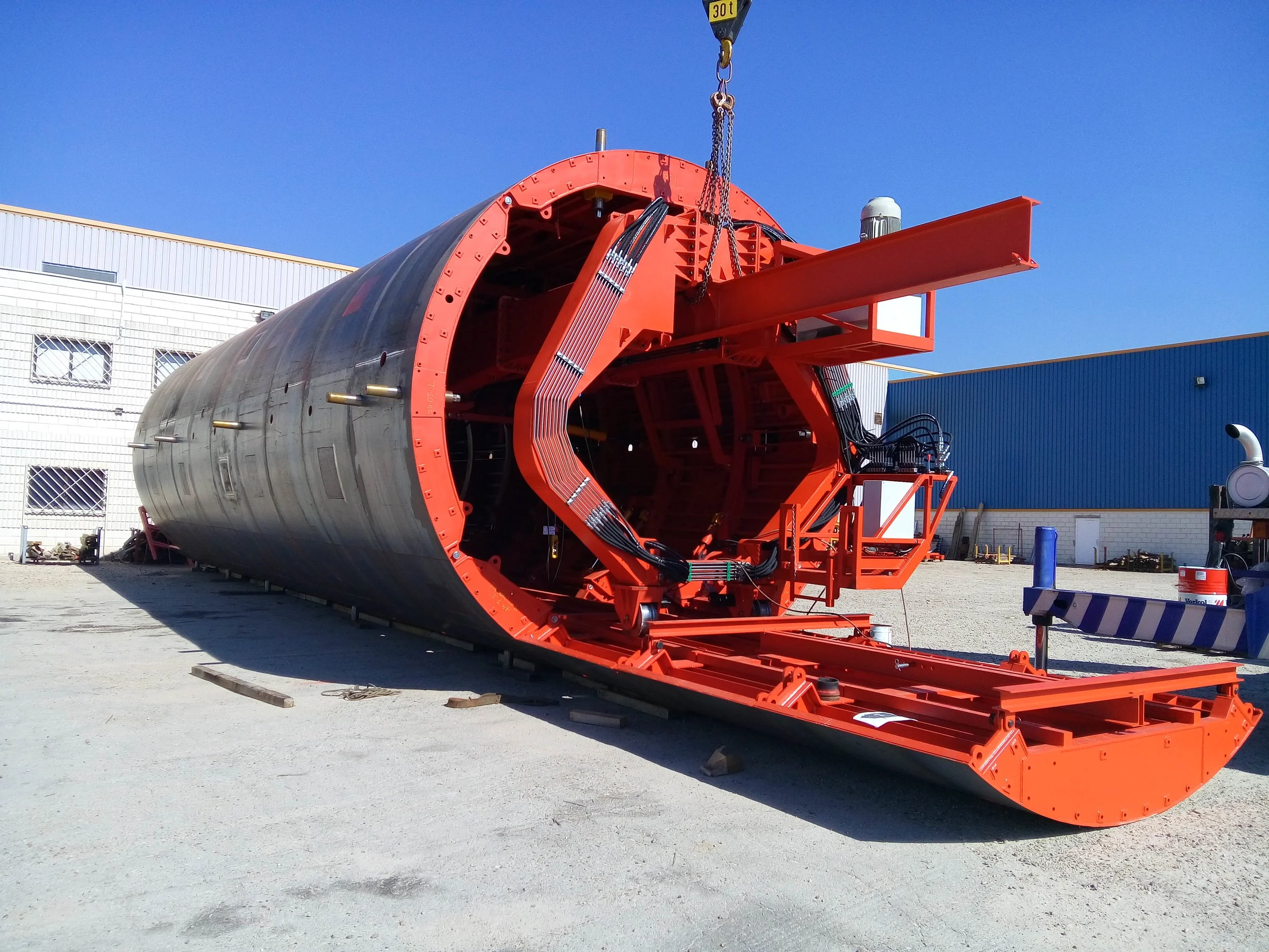 Large industrial machine with a cylindrical body and orange structural supports, placed outdoors near industrial buildings.