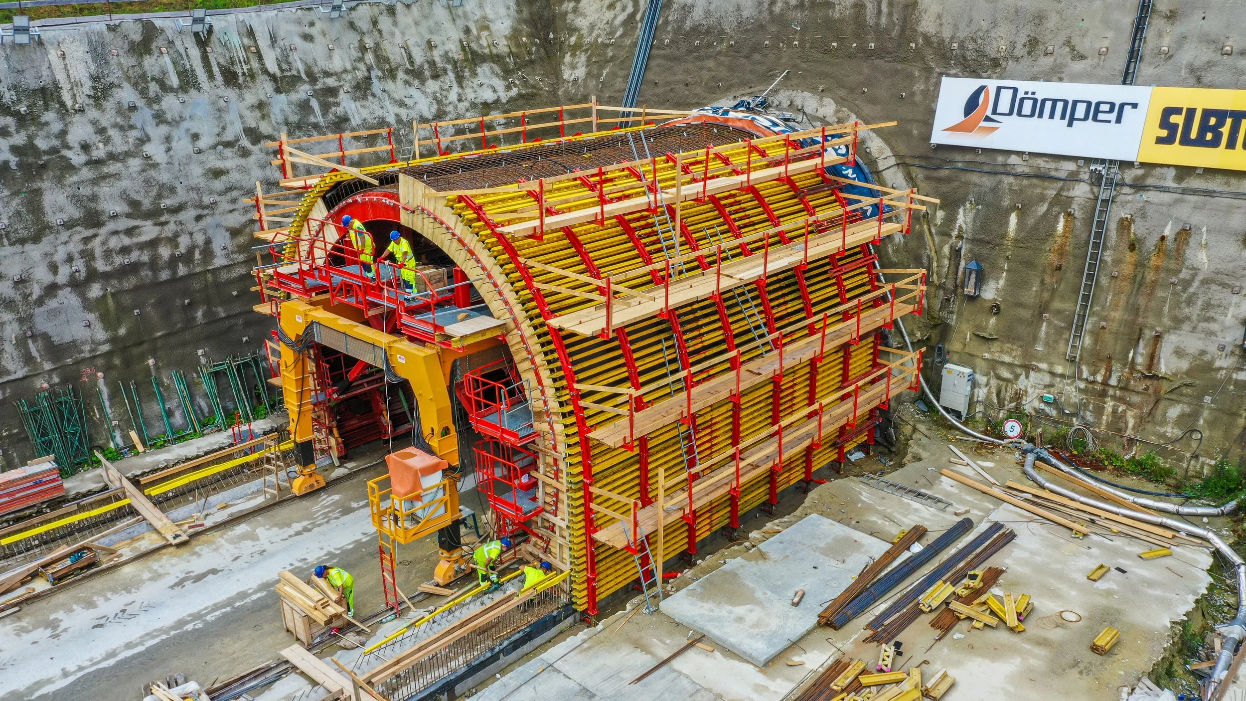 Construction site with workers building a tunnel using a large, semi-circular shape structure supported by red and yellow framework. Construction materials and equipment are scattered around the site, and a sign with some logos is visible on the wall