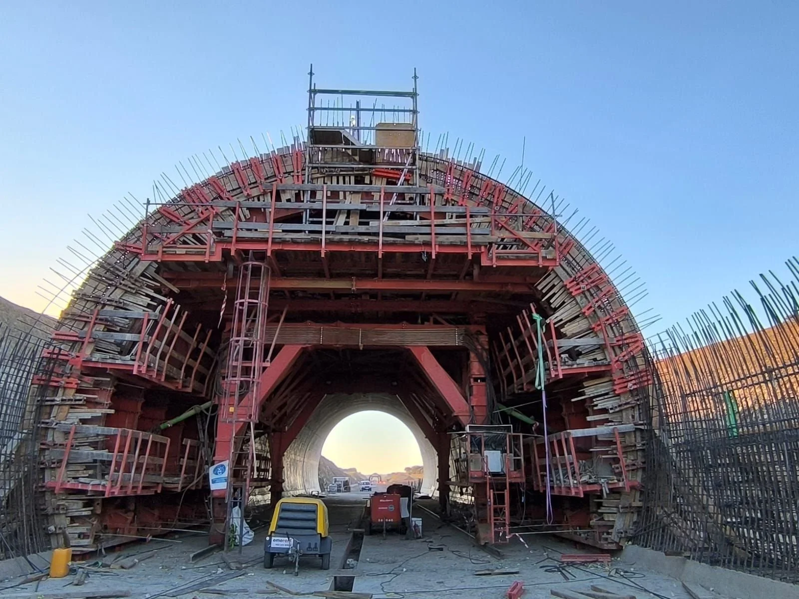 Under construction tunnel with scaffolding and safety barriers, trucks, and construction equipment on fresh construction site.