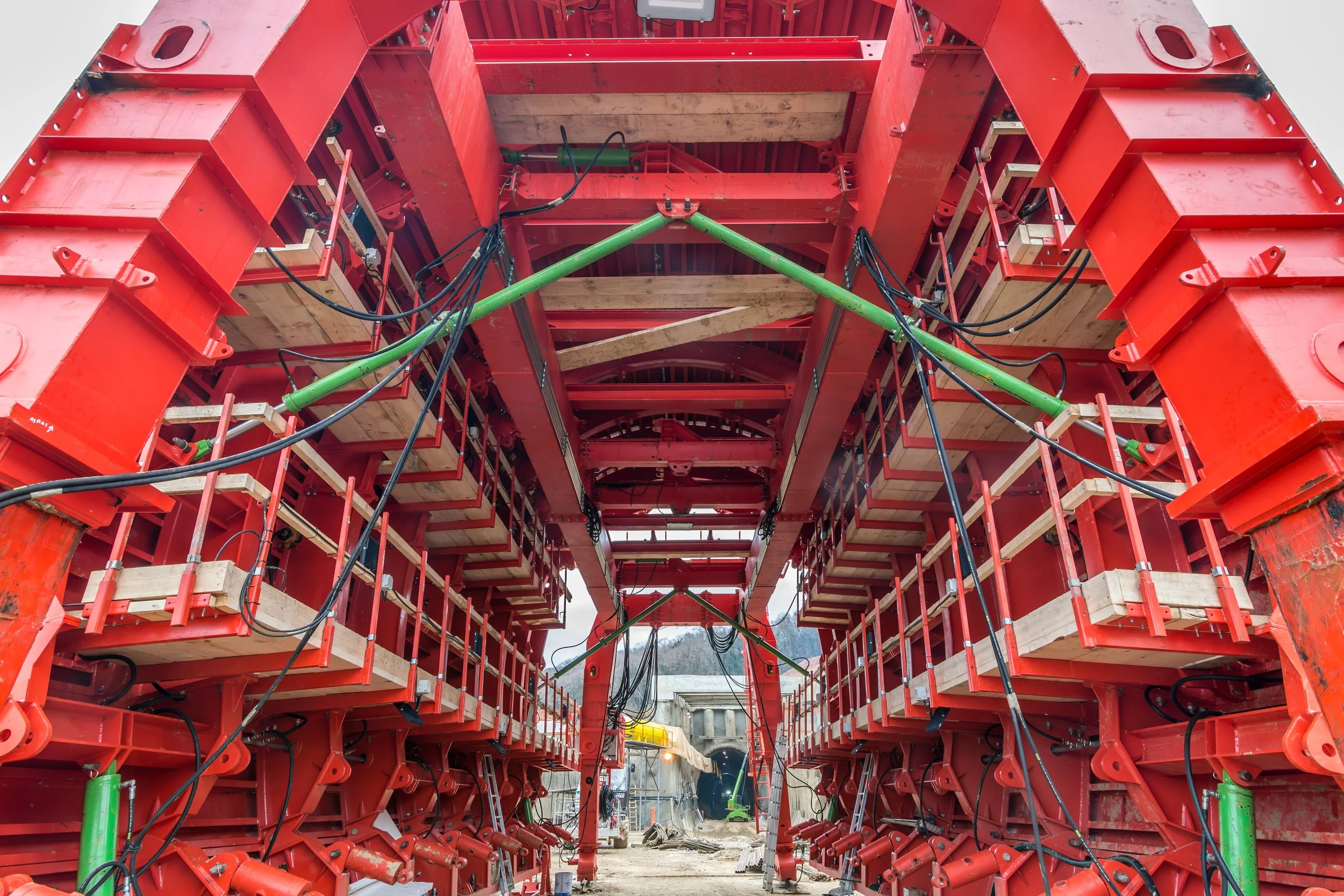 Construction site with large red machinery and green hydraulic components leading towards a tunnel in the background.