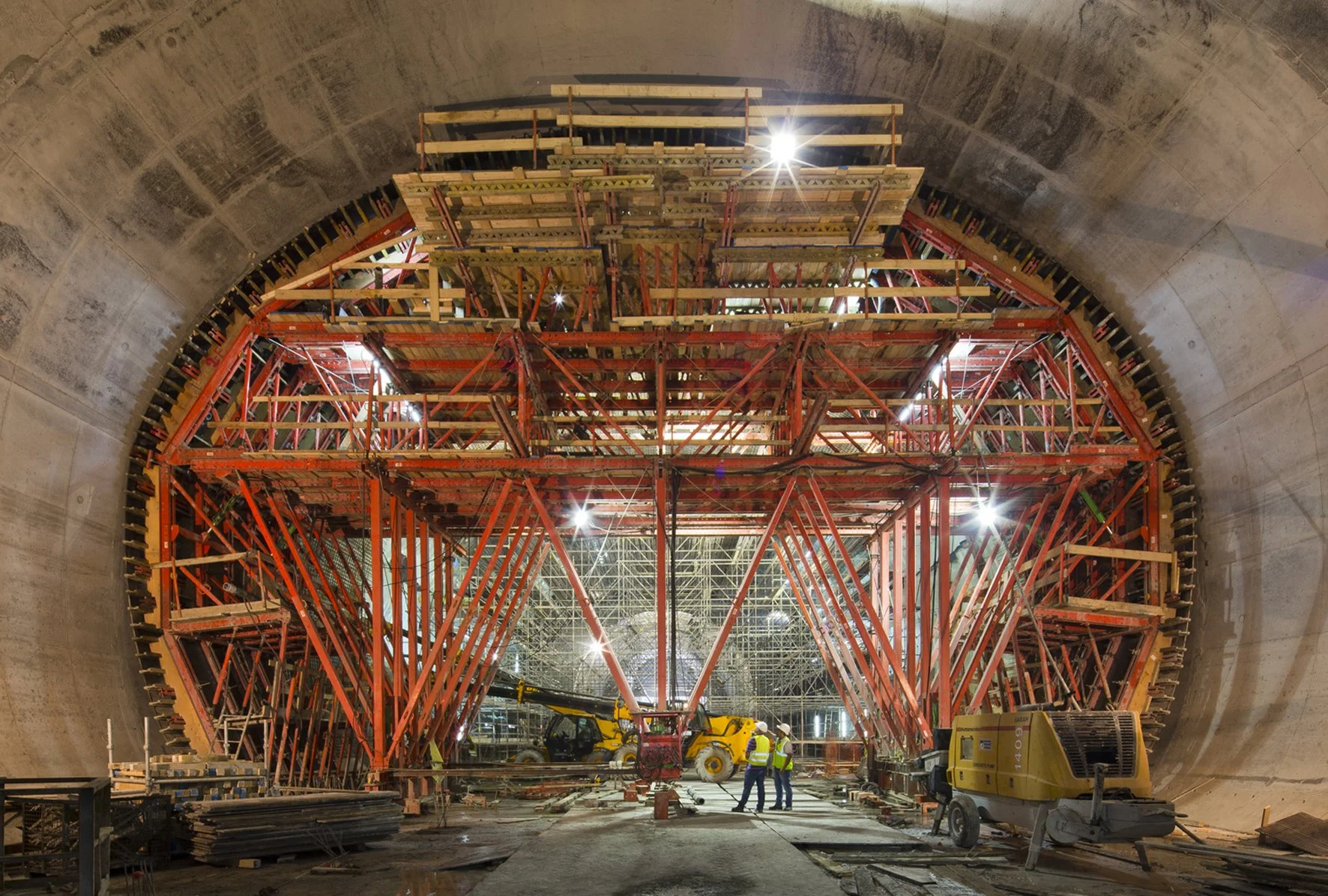Construction workers and machinery inside a tunnel under construction, with intricate scaffolding and red structural supports.