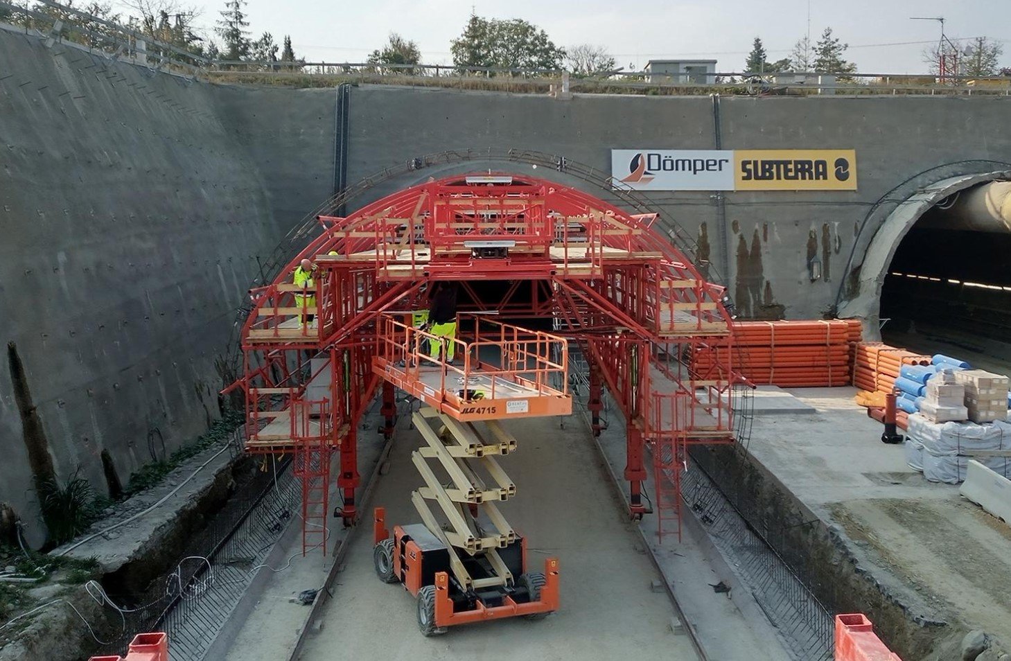 Construction workers in high-visibility clothing working on a tunnel under construction, with scaffolding and equipment around the entrance.