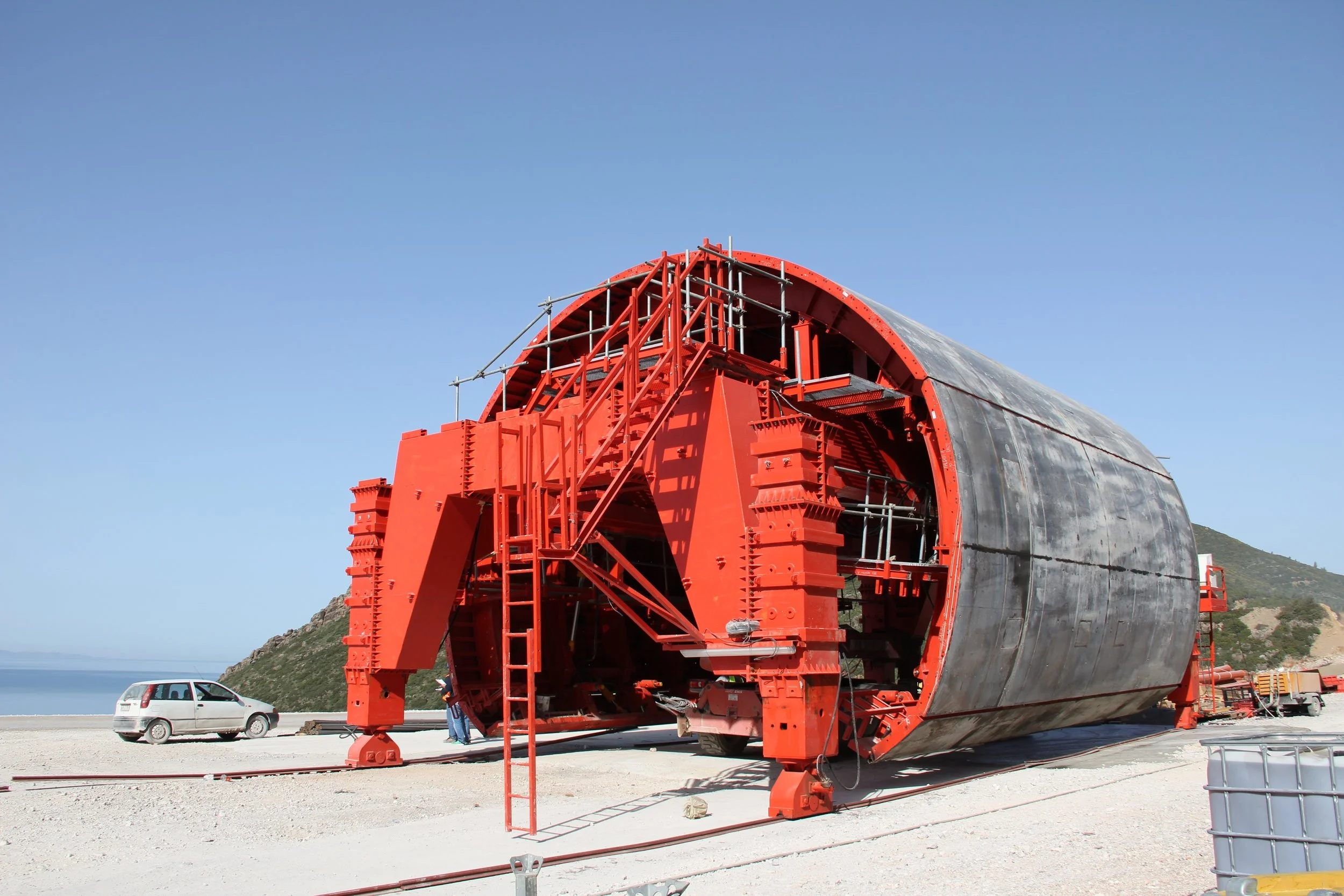 A large, cylindrical, red industrial structure on a sandy area near a coastal landscape under a clear blue sky.