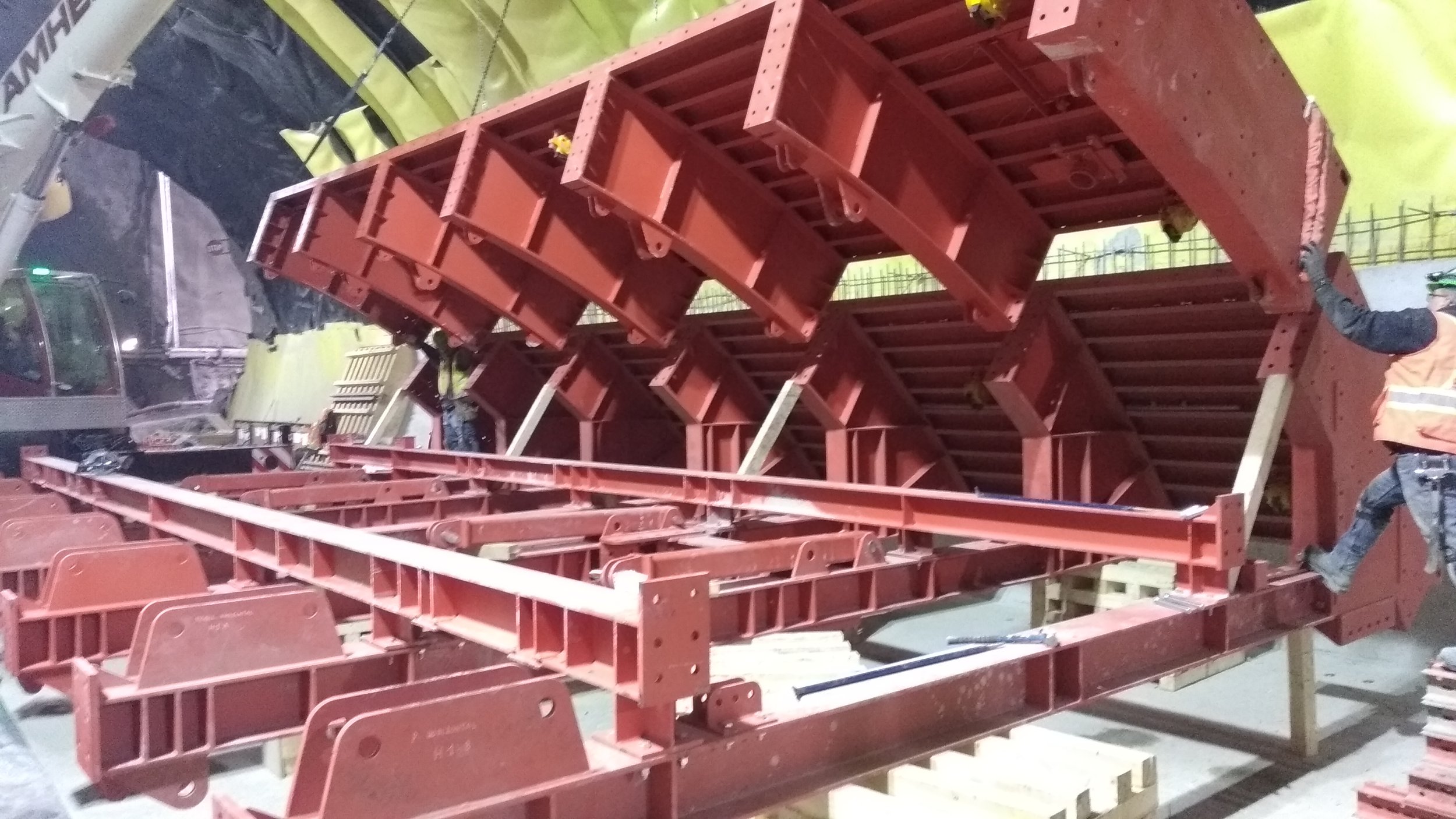 Large red steel framework structure under construction at a construction site, with workers and construction equipment visible.