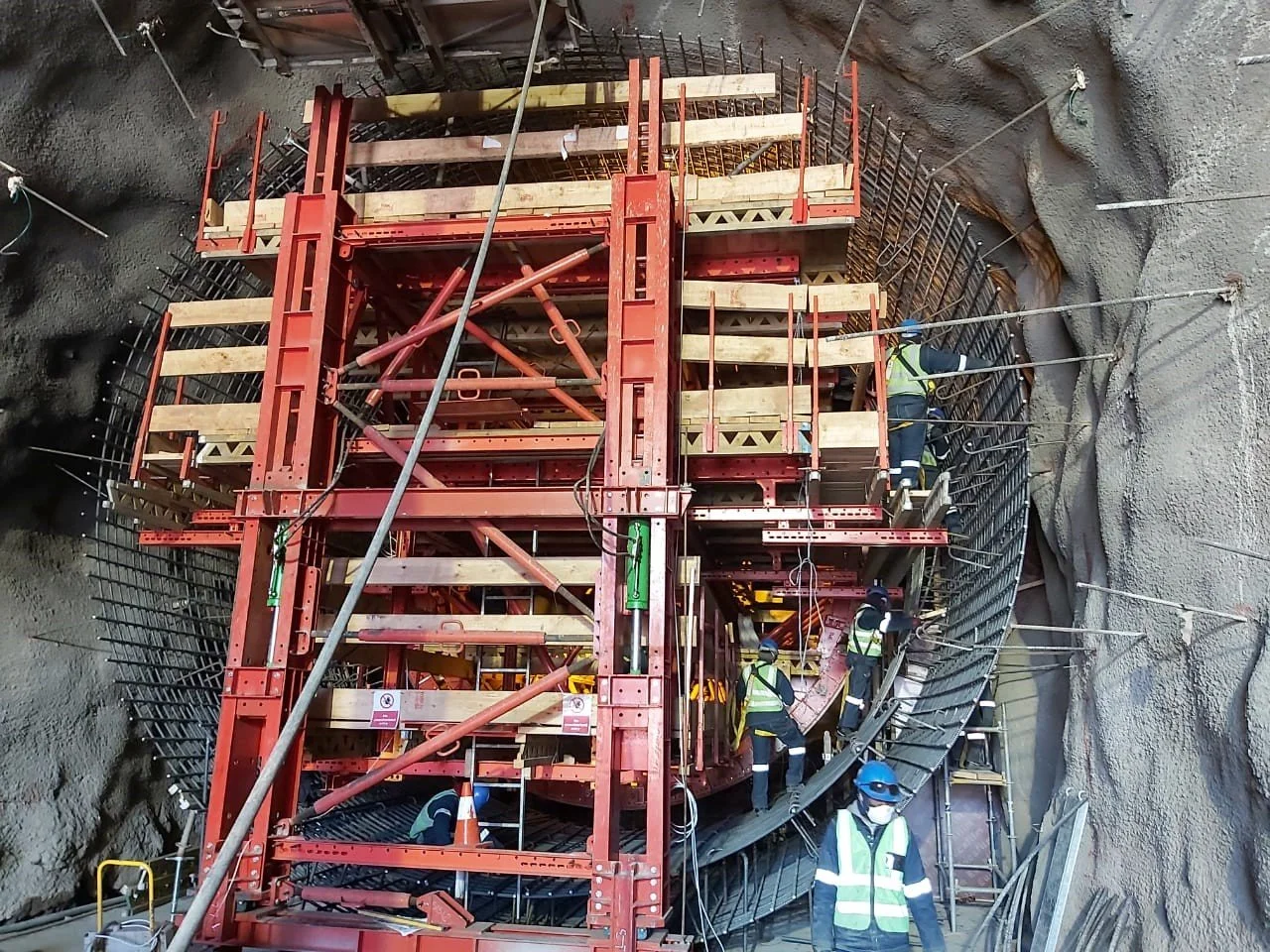 Construction workers assembling scaffolding inside a deep circular tunnel with steel rebar and concrete walls.