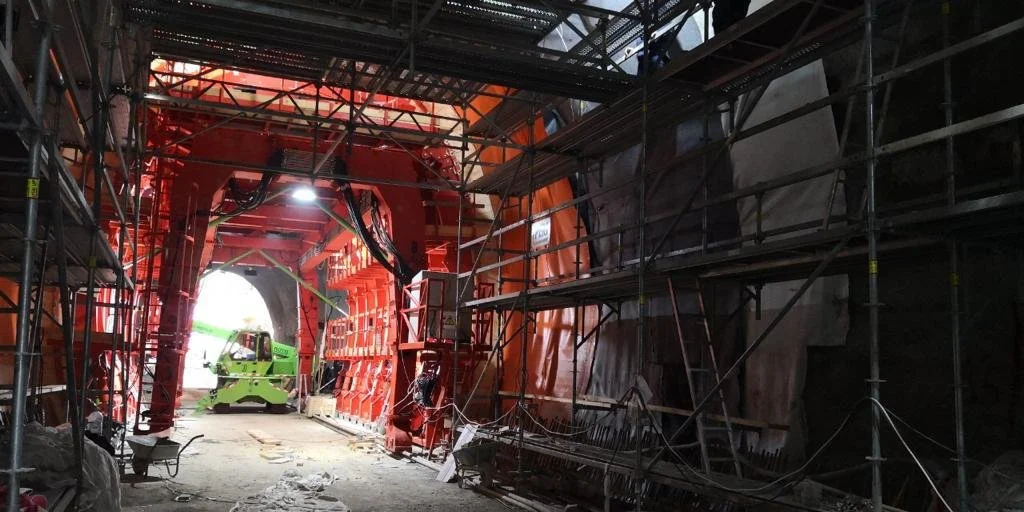 Construction site inside a tunnel with scaffolding and machinery, including a green lift, and bright light at the tunnel's exit.