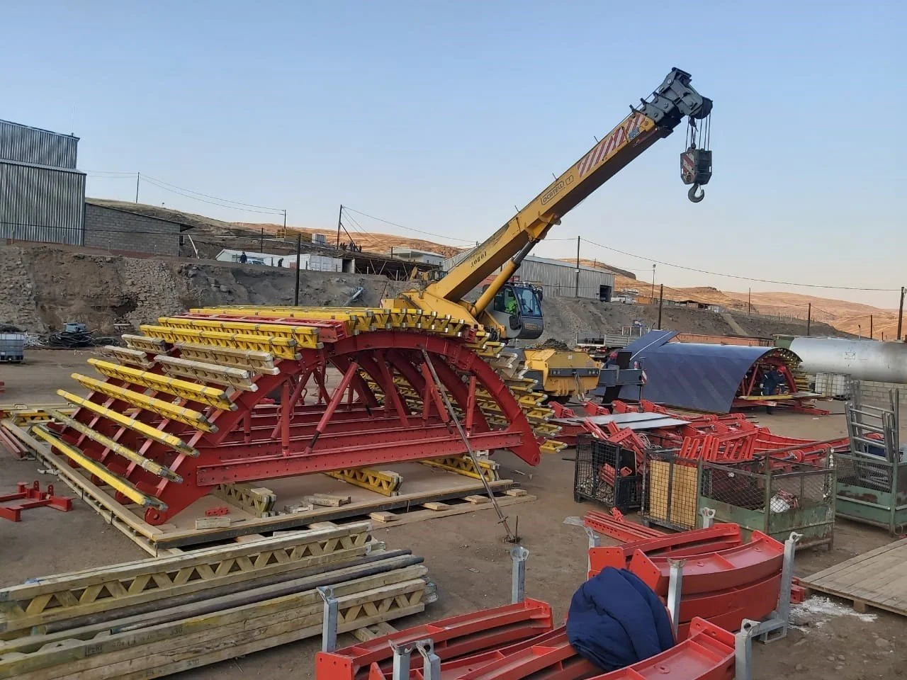 Construction site with large red and yellow machinery, including a crane, pipes, and building materials in a rural area.