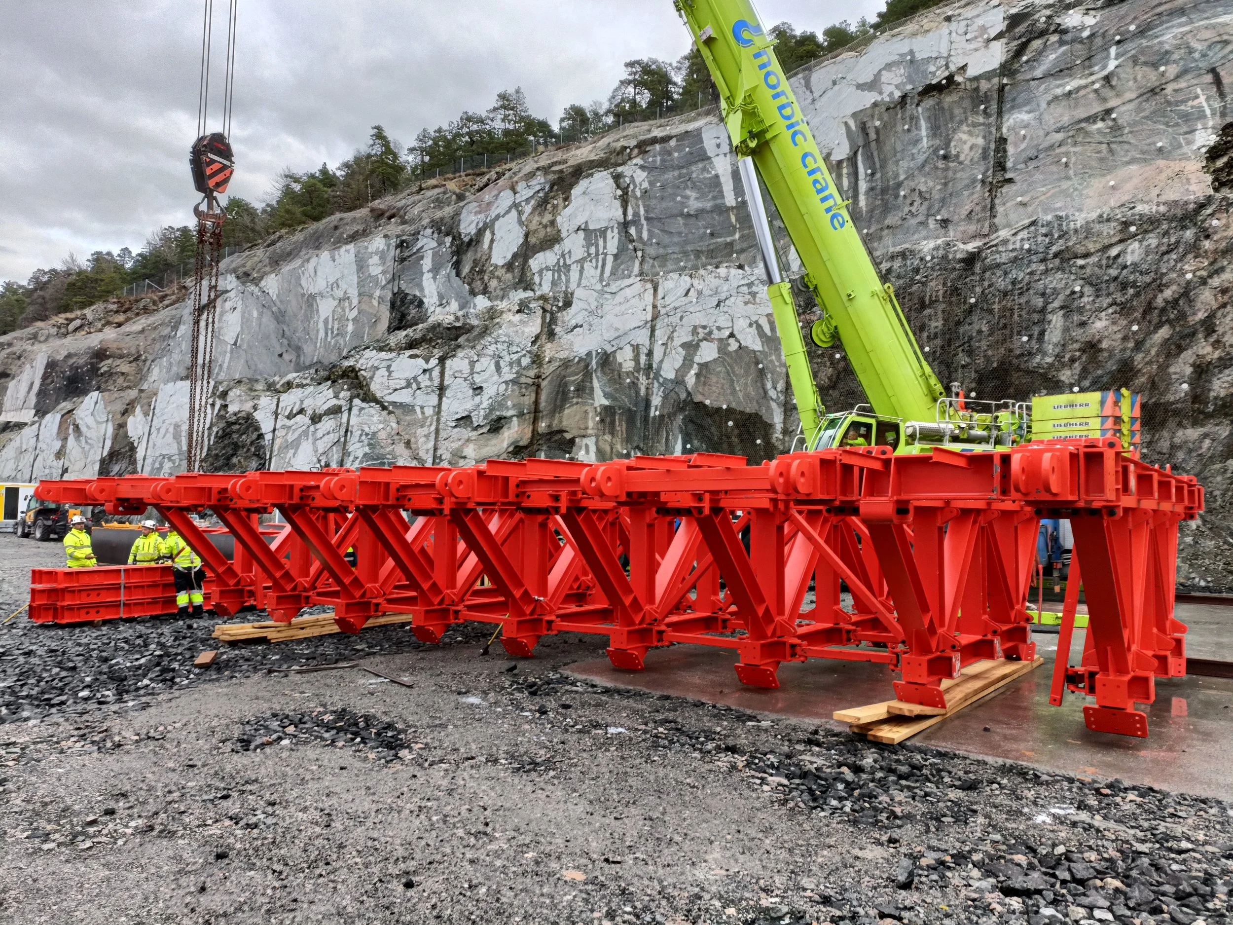 Construction site with a large green crane lifting a red modular structure against a rocky cliff. Three workers in yellow safety gear are standing nearby.