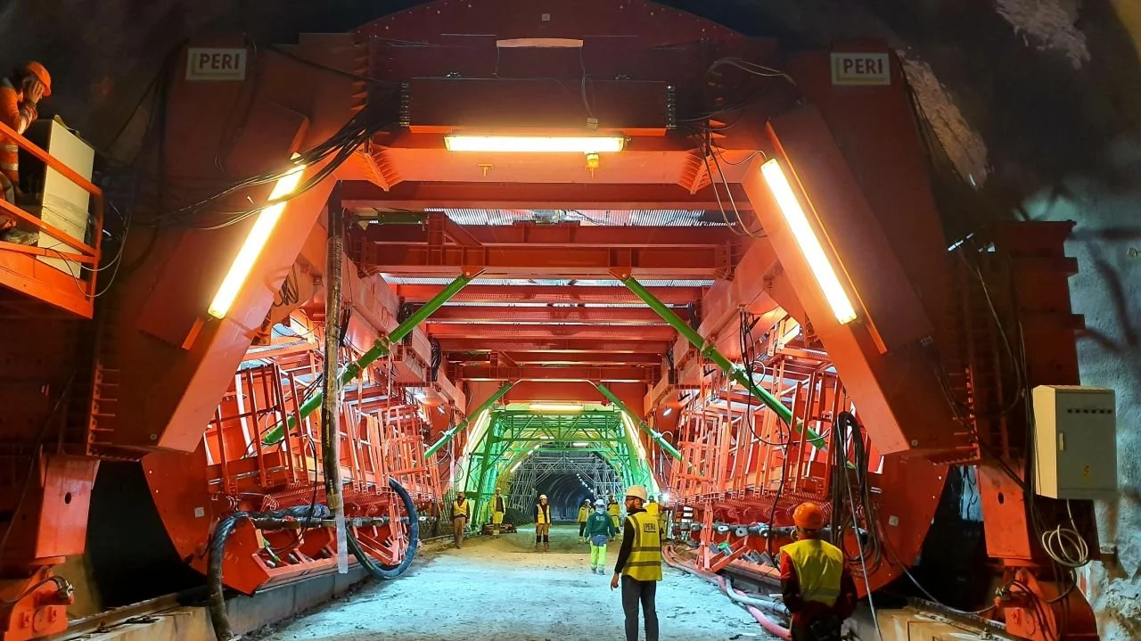 Workers inside a large tunnel construction site, wearing safety vests and helmets, with massive machinery and lighting installed in the tunnel walls.