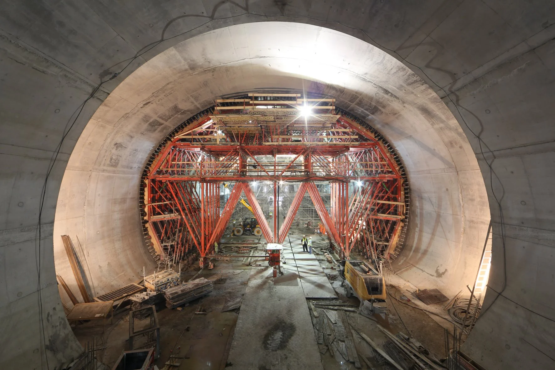 Underground construction site with large tunnel and red scaffolding inside the tunnel, construction workers, and equipment.