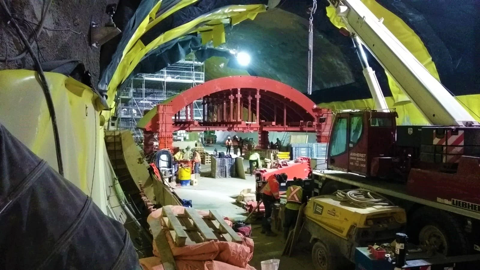 Construction workers working inside an underground tunnel with a red structural arch, scaffolding, and construction equipment.