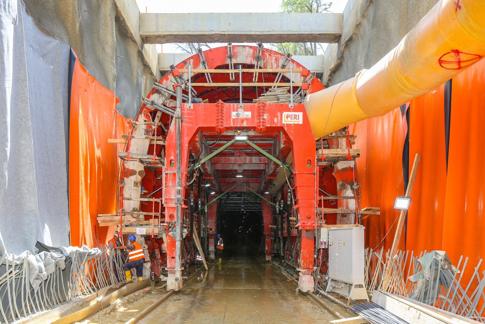Construction workers in safety vests and helmets working inside tunnel construction site with large orange and yellow machinery and tunnel lining.