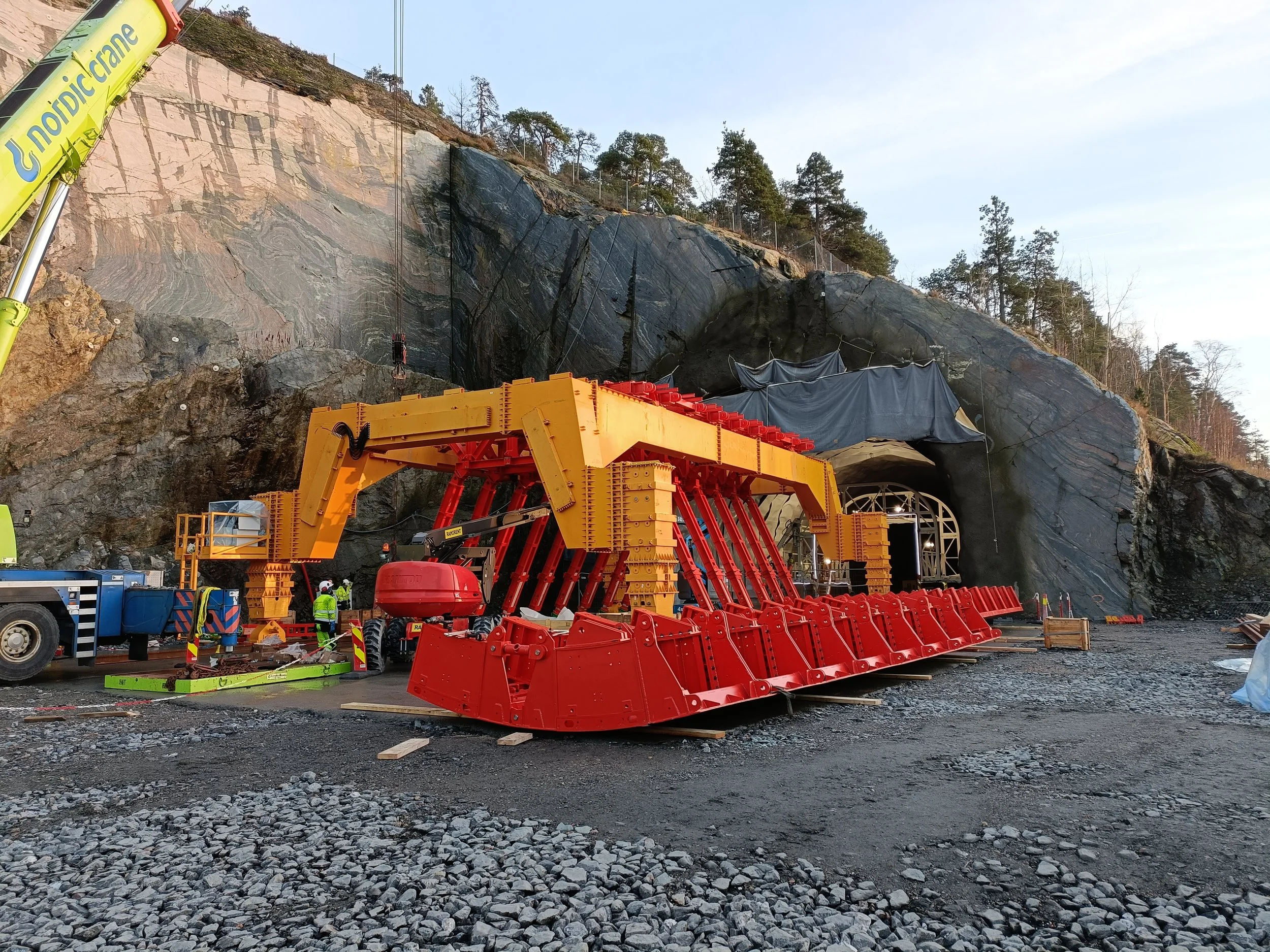 A construction site at the entrance of a tunnel through a rocky hillside, with heavy machinery including a large yellow and orange tunnel boring machine and workers wearing safety gear.