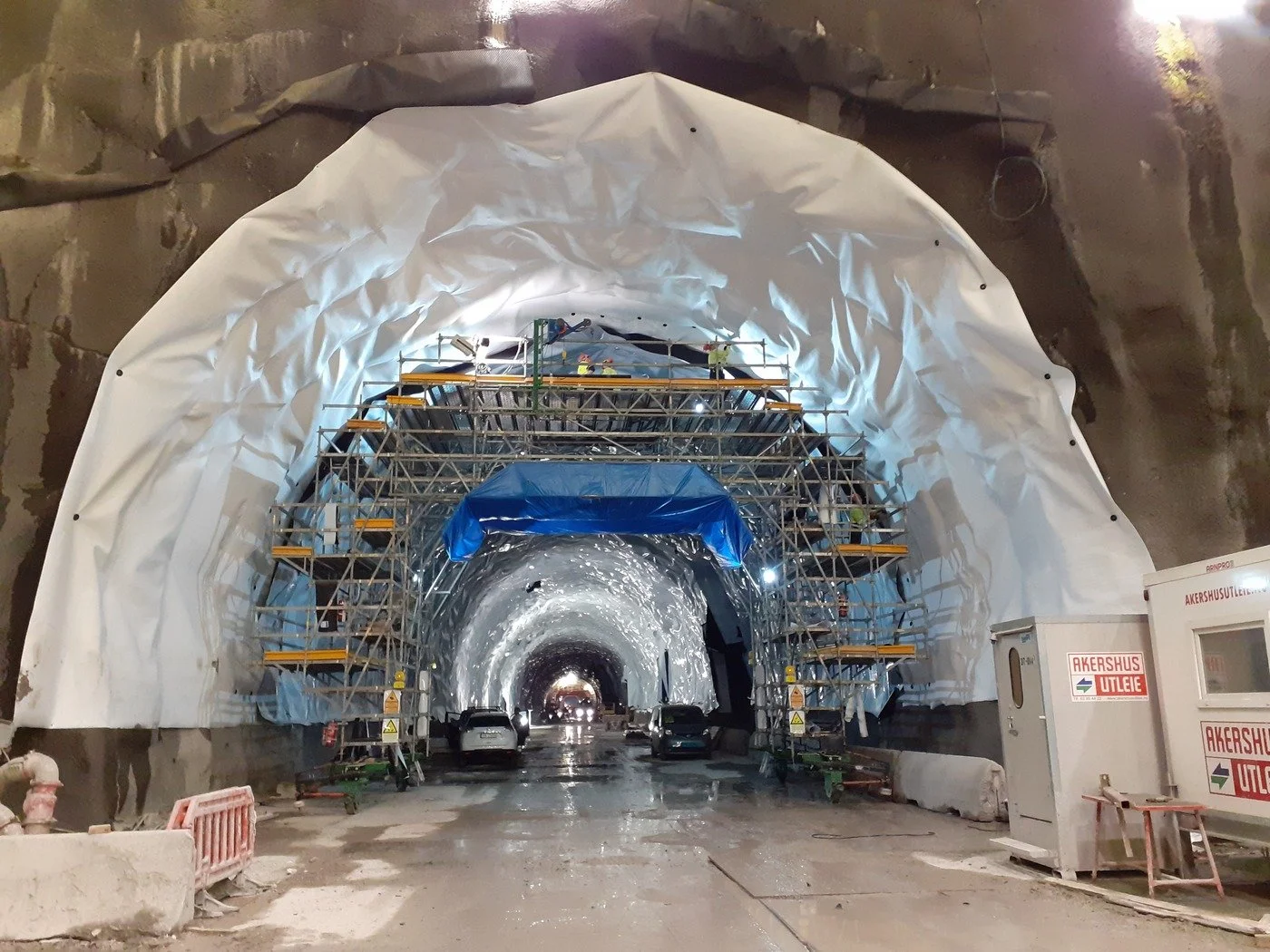 Construction workers working on scaffolding inside a tunnel under construction, with cars parked and construction equipment around.