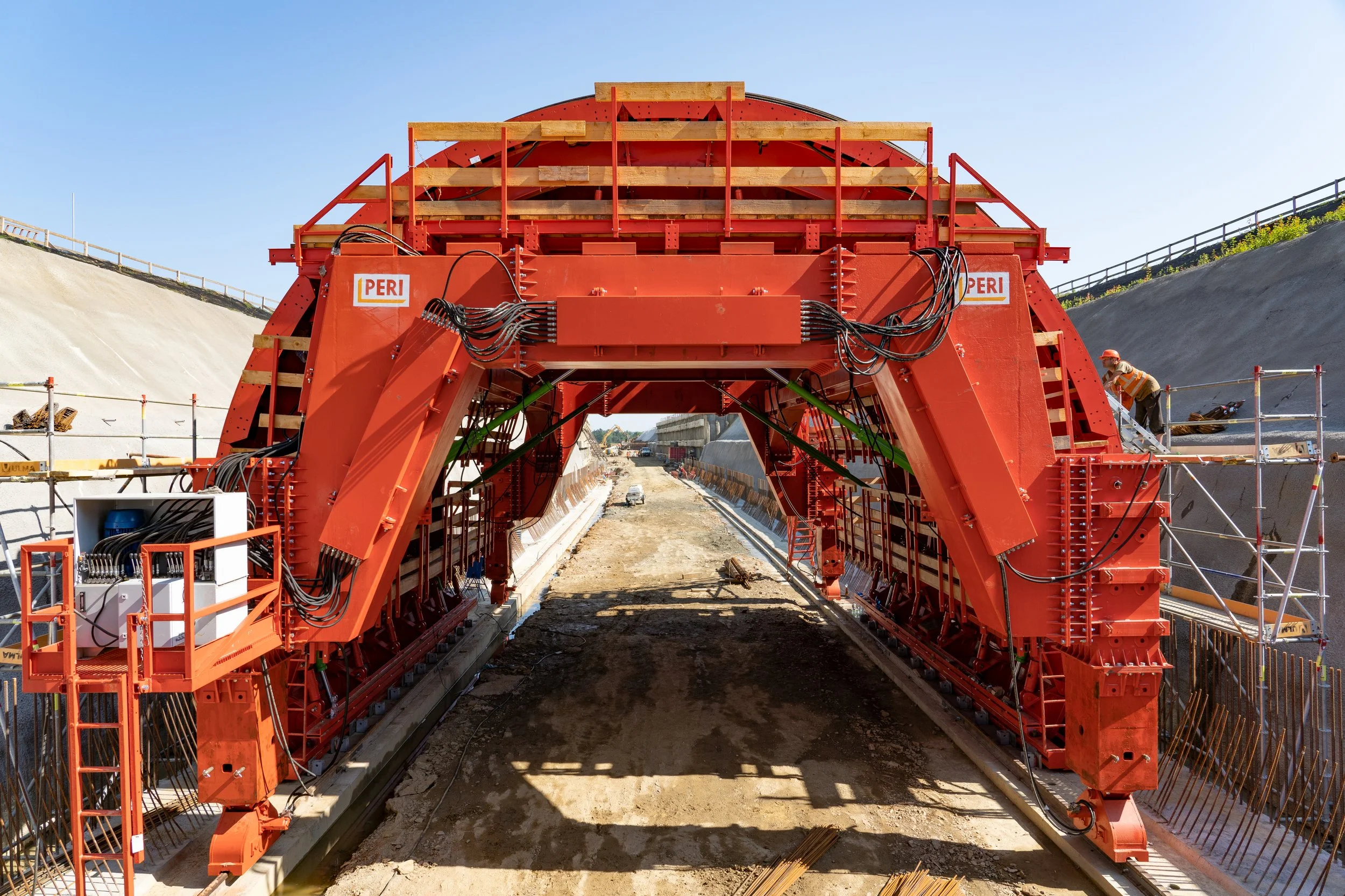 A large red tunnel boring machine at a construction site with workers working on the sides, and a clear sky overhead.