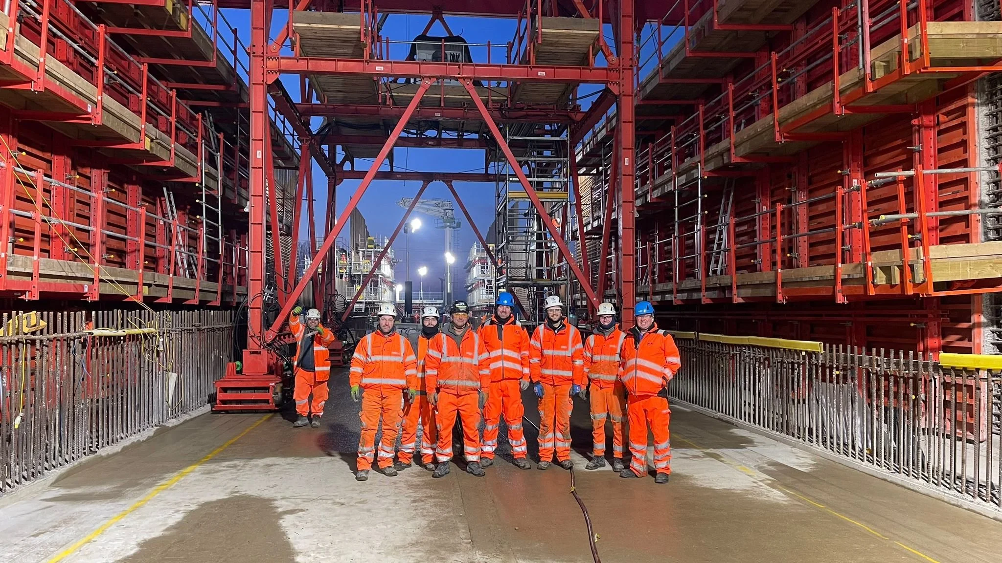 Group of construction workers in orange safety gear and helmets standing on a construction site with large red scaffolding and equipment.