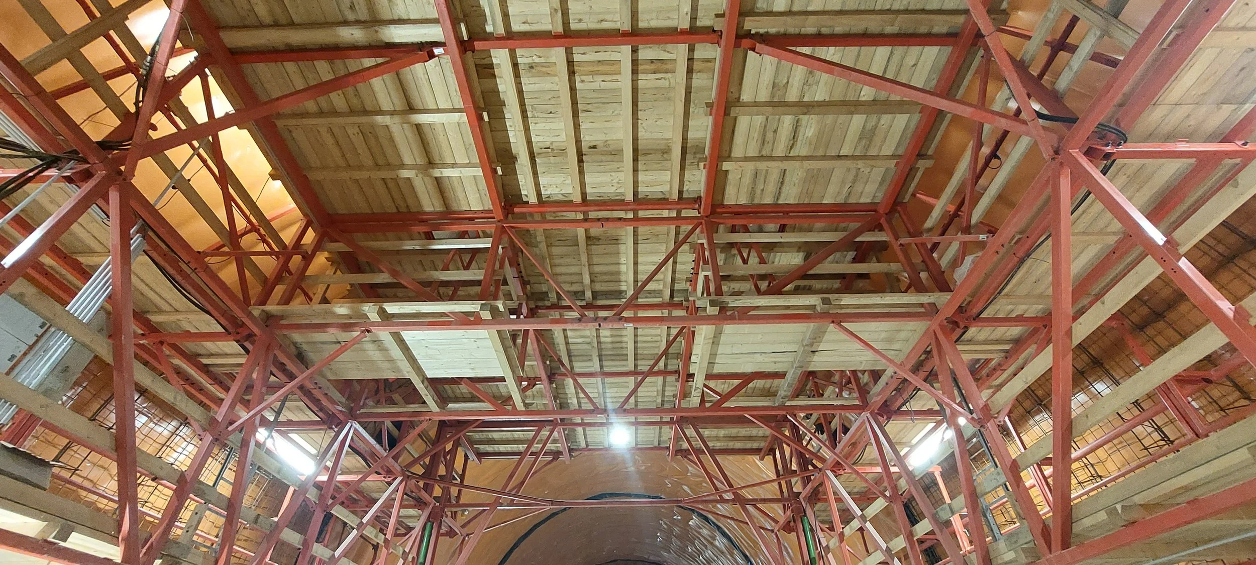 Construction site roof with red scaffolding and wooden ceiling beams.
