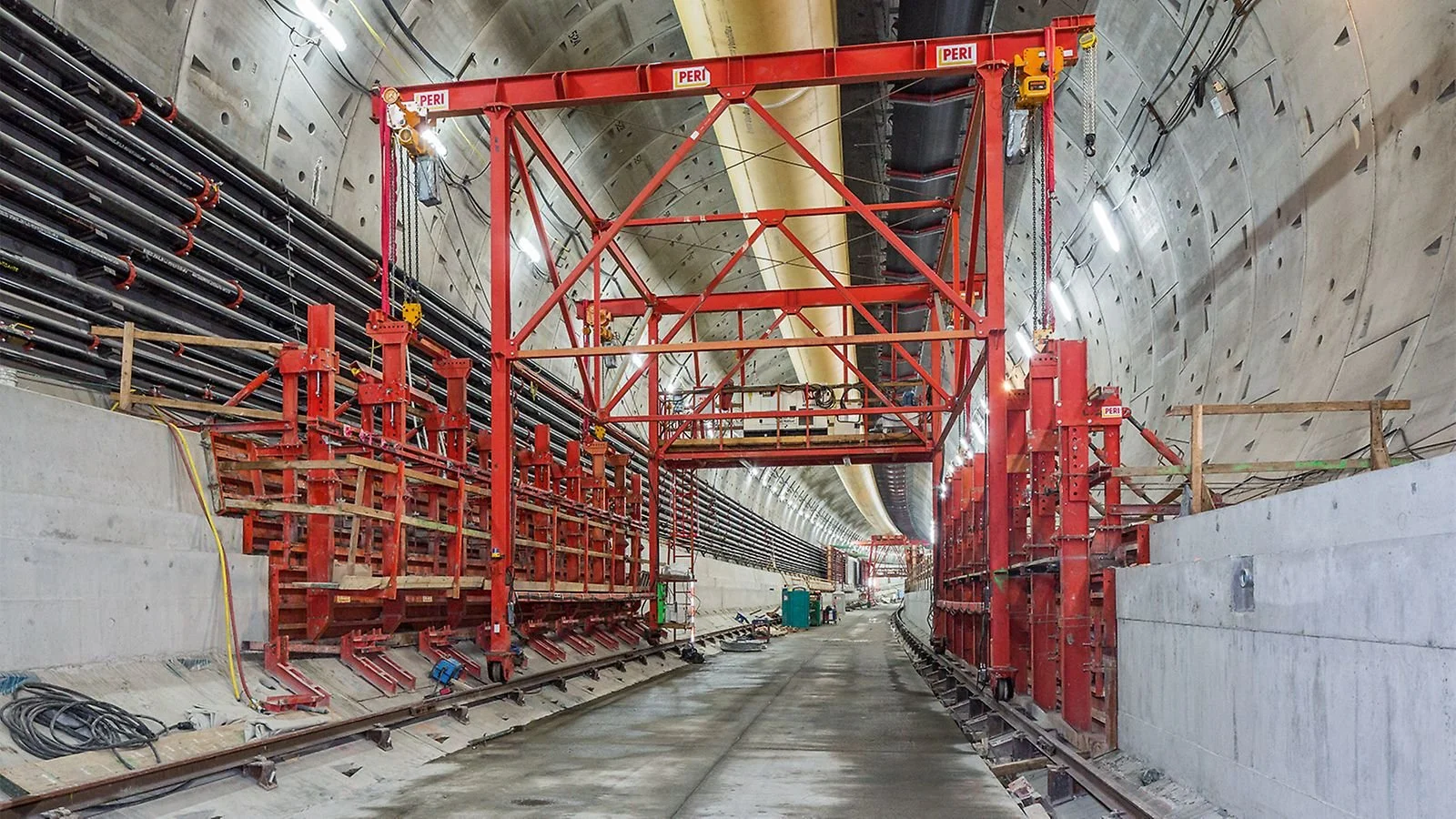 Construction inside a tunnel featuring red scaffolding used for tunnel boring operations, with pipelines and construction equipment along the sides.