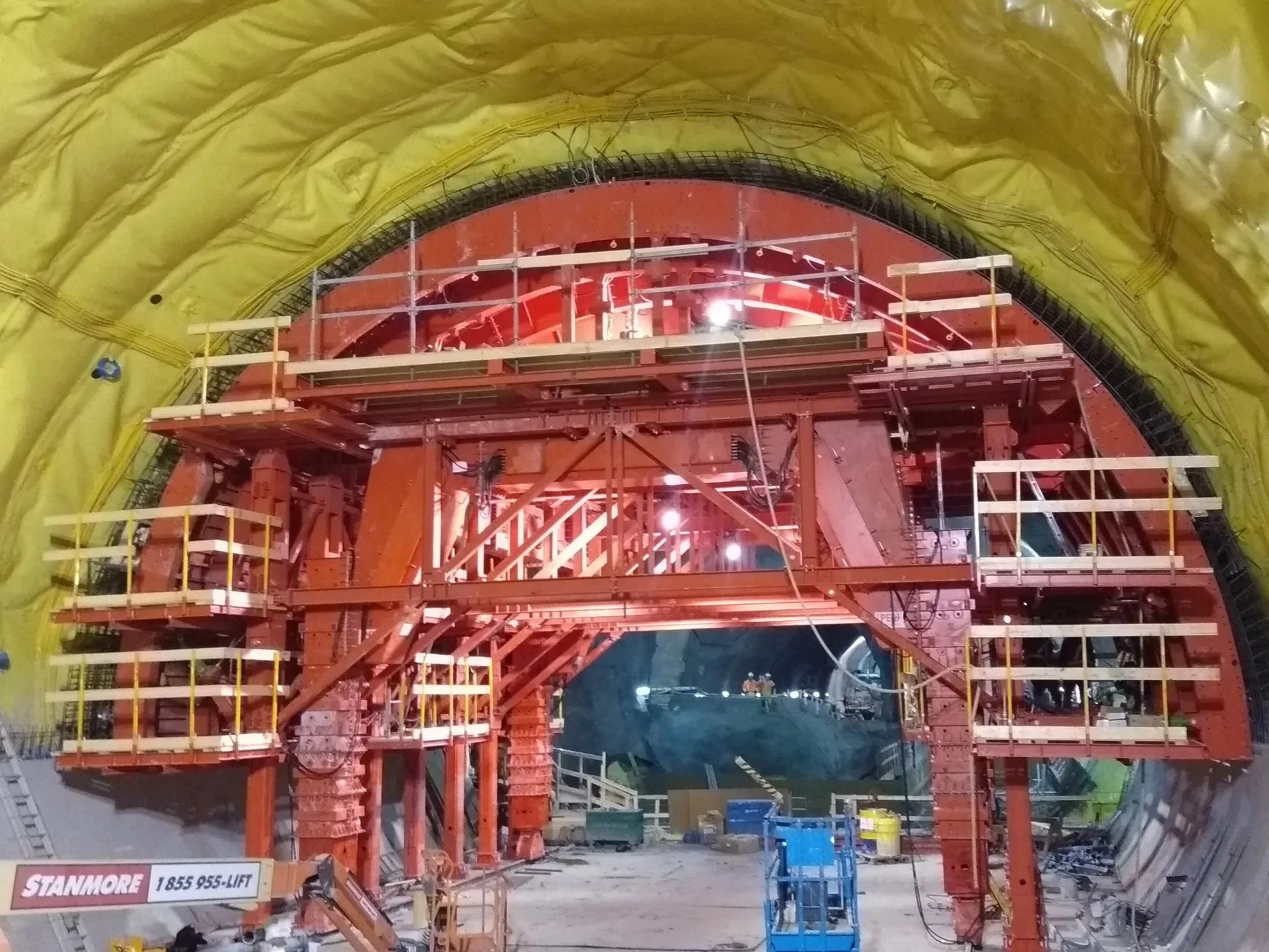 Construction workers standing inside a large tunnel bore machine with yellow lining, red structural components, and scaffolding.